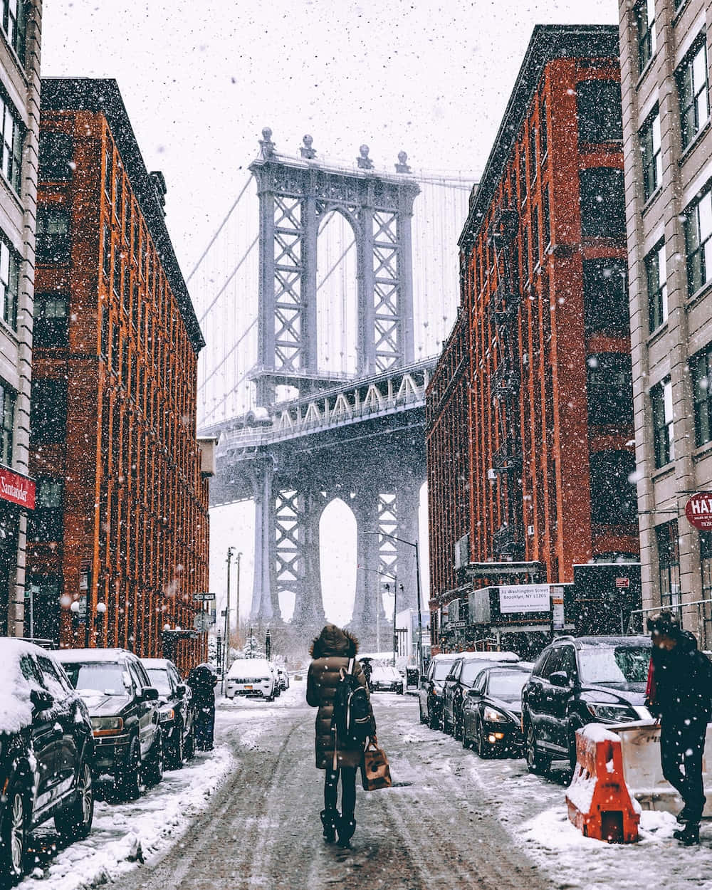 雪 桌布 壁紙 — 一個孤獨的身影在白雪覆蓋的城市街道上行走，兩旁是磚砌建築，曼哈頓大橋在輕柔的降雪中高聳入雲。