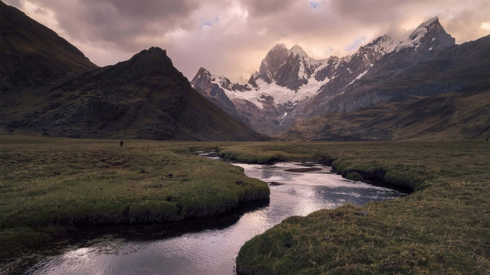 Beautiful Landscape 桌布 壁紙 — 一個孤獨的身影在蜿蜒的河流旁的草地上行走，背景是戲劇性的多雲天空和遠處的雪山。