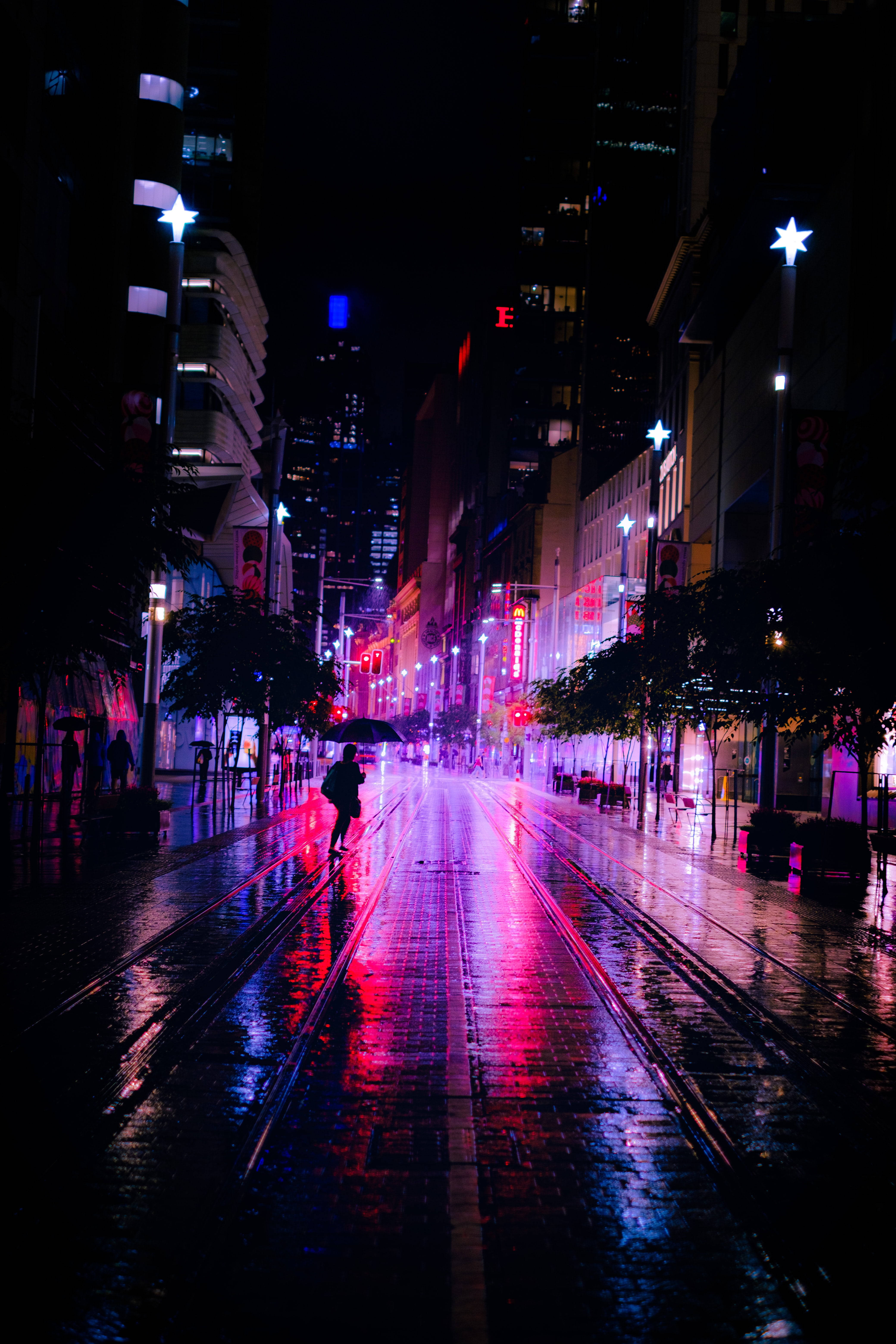 Dark Neon 桌布 壁紙 — 一個孤獨的身影在雨夜中撐著傘，沿著霓虹燈閃爍的濕滑城市街道行走，發光的星形路燈和濕漉漉路面上的倒影清晰可見。