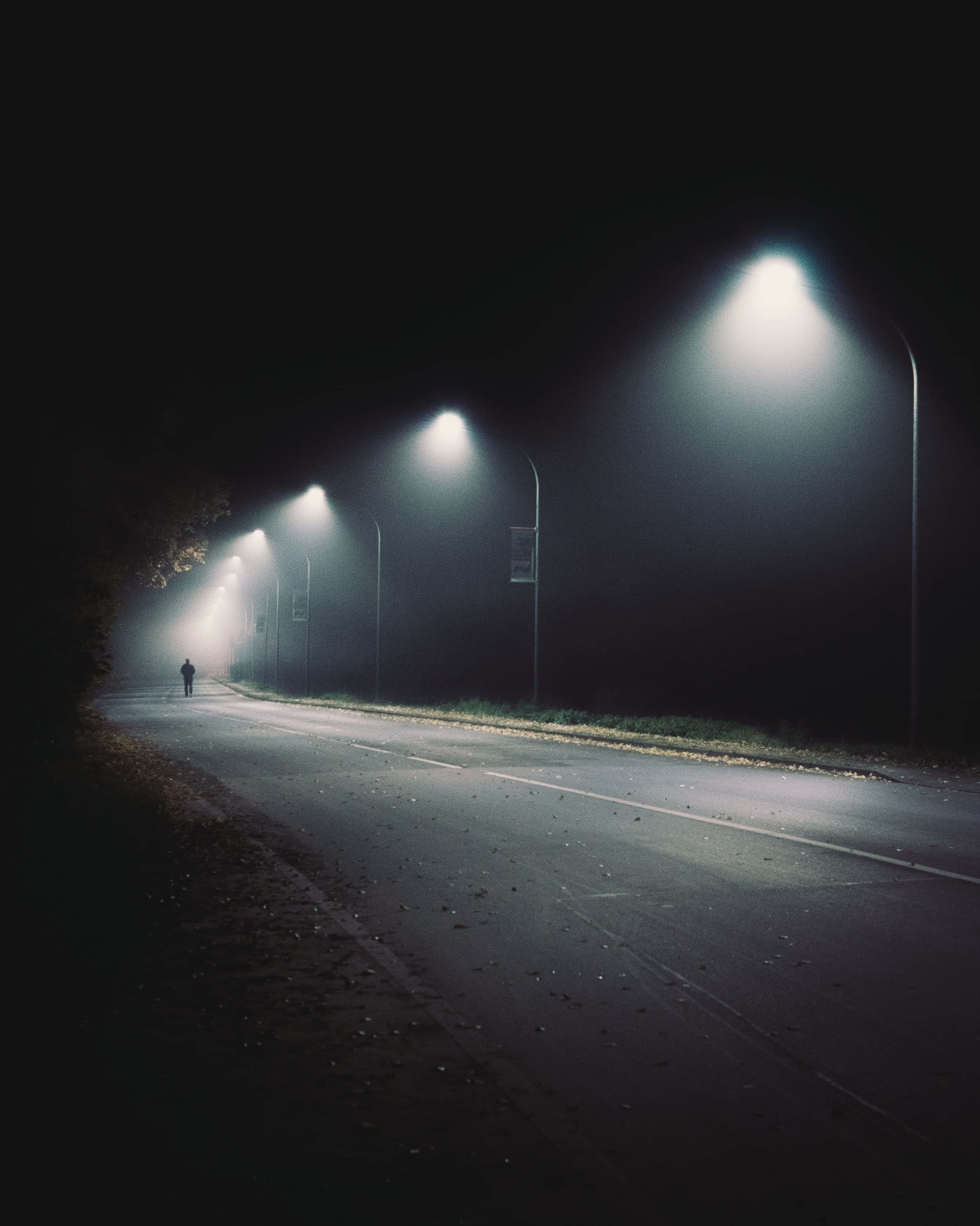 Night Time 桌布 壁紙 — 一個孤獨的身影在霧氣瀰漫的夜路上行走，路燈成排，在濕漉漉的瀝青上投下柔和的光芒。