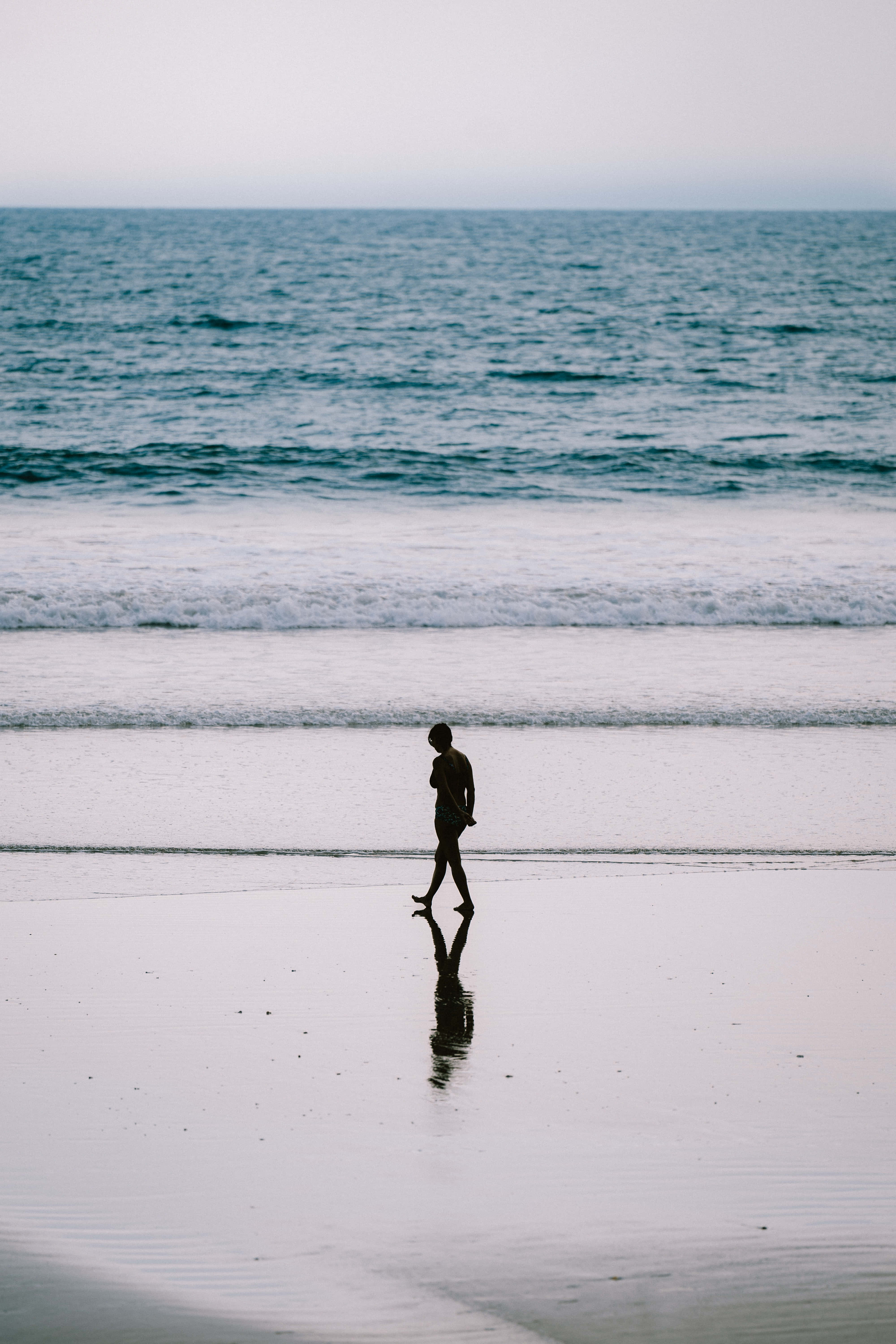 Aesthetic Beachy 桌布 壁紙 — 一個孤獨的身影在黃昏時沿著濕漉漉的海灘行走，身後有海浪翻滾。