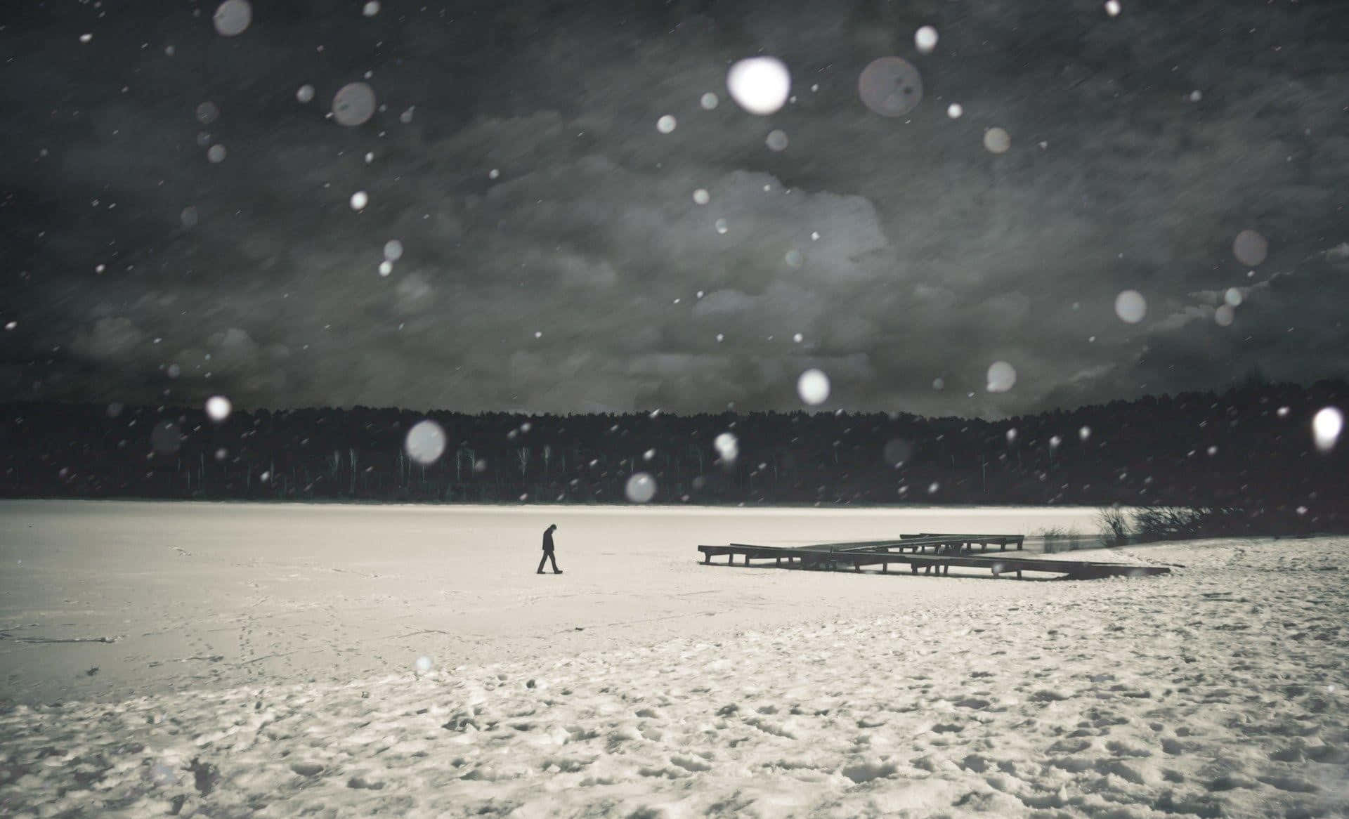 Lonely 桌布 壁紙 — 一個孤獨的身影在黑暗暴風雪的天空下，走過白雪覆蓋的冰凍湖面，雪花飄落，散景燈光閃爍。