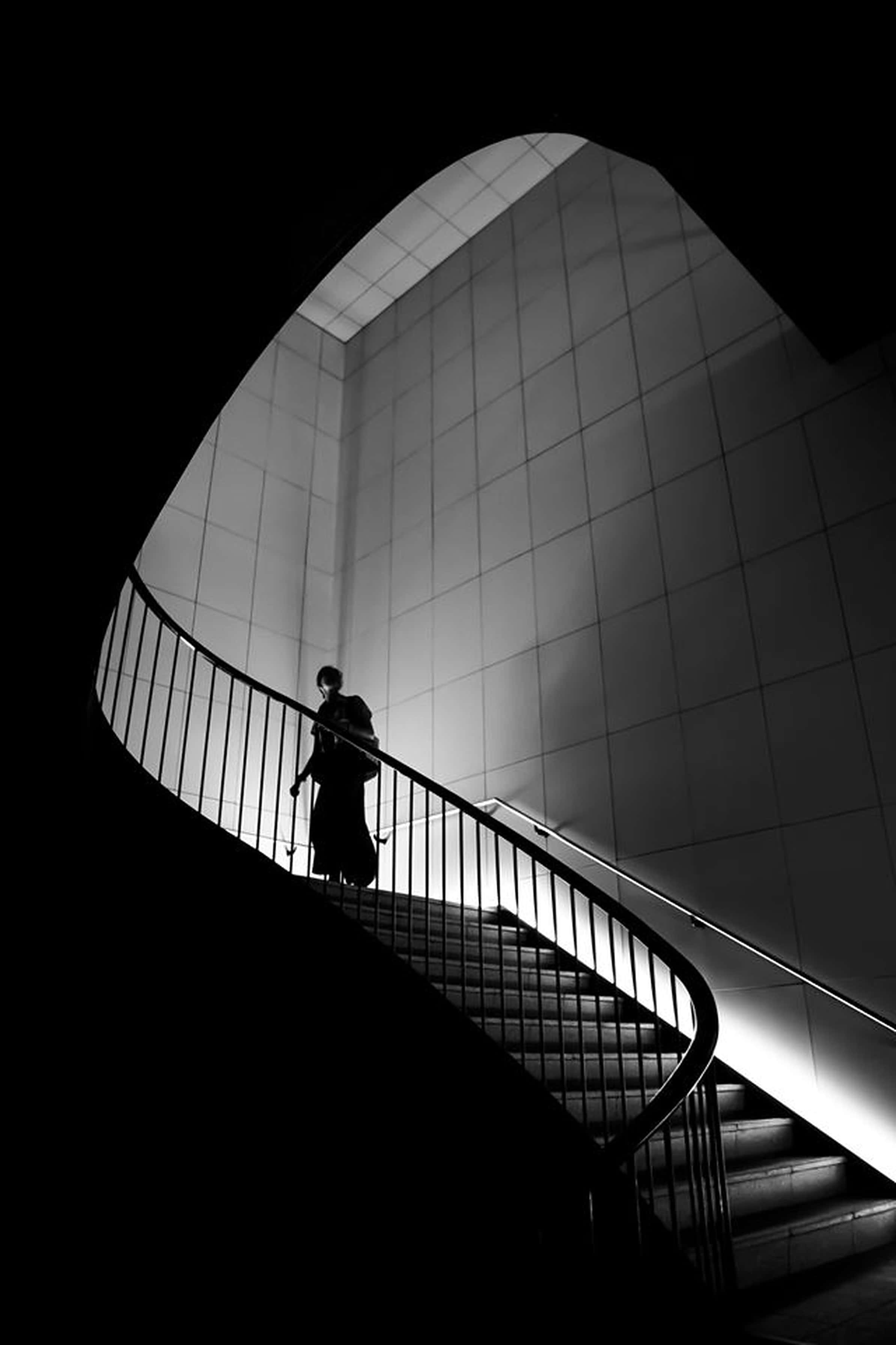 Cool Black And White 桌布 壁紙 — 一個孤獨的身影在黑白建築空間中，沿著戲劇性照明的螺旋樓梯向上走。