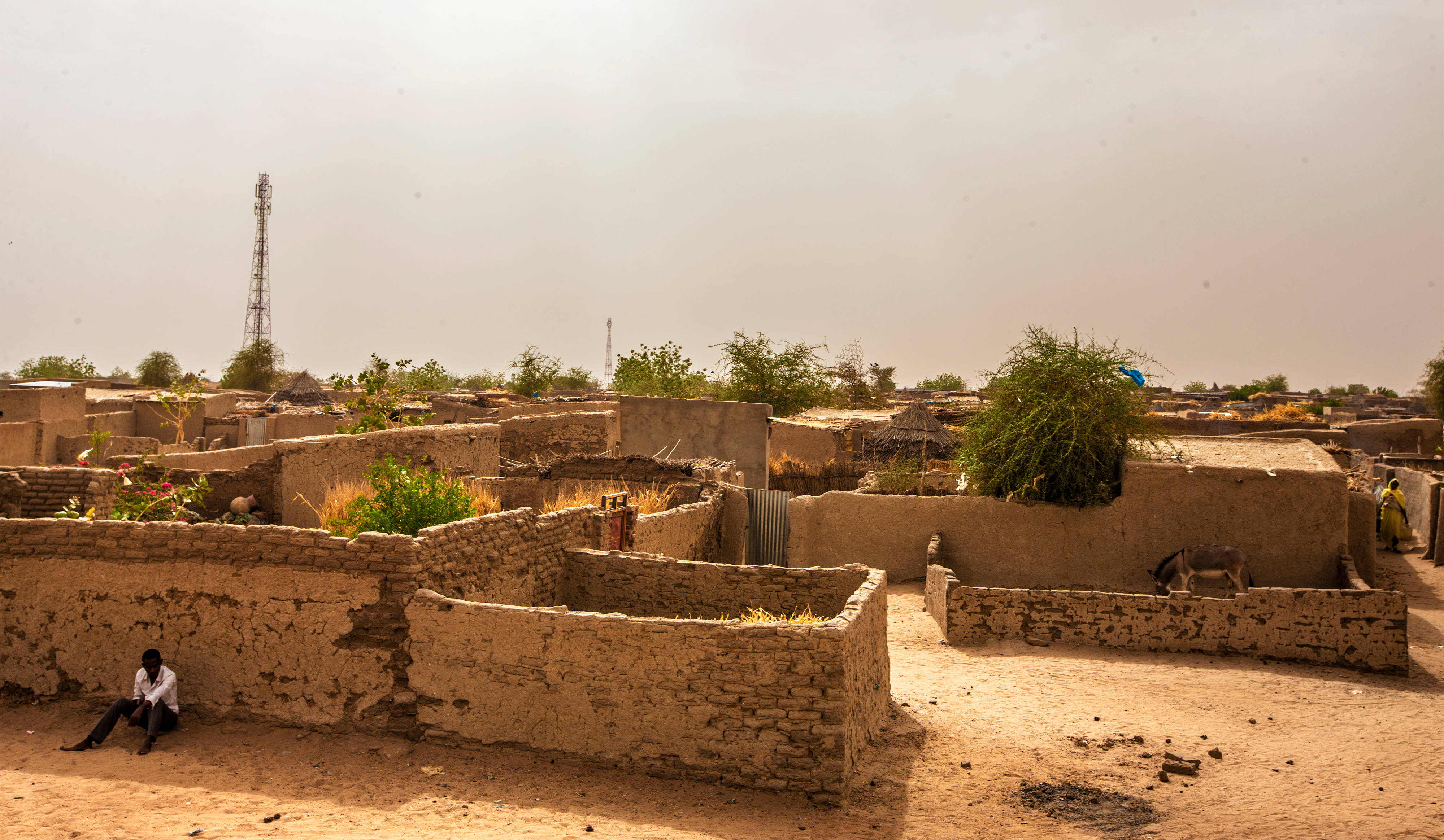 Sudan 桌布 壁紙 — 一個孤獨的身影坐在塵土飛揚的非洲村莊裡，靠著被太陽曬乾的泥磚牆，天空朦朧。