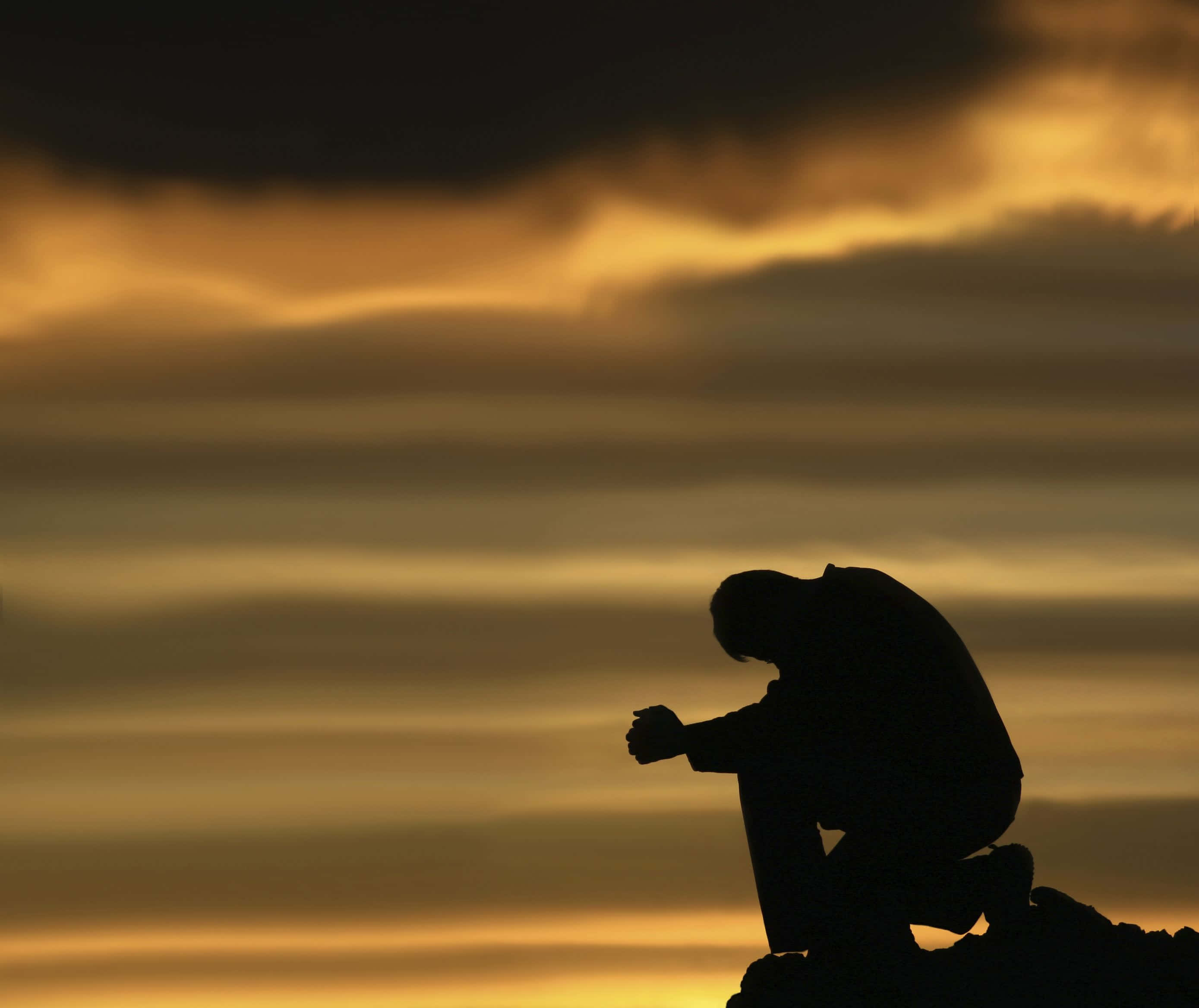 Praying 桌布 壁紙 — 一個孤獨的身影坐在岩石上，剪影映襯著佈滿金色琥珀色雲彩的壯麗日落天空。