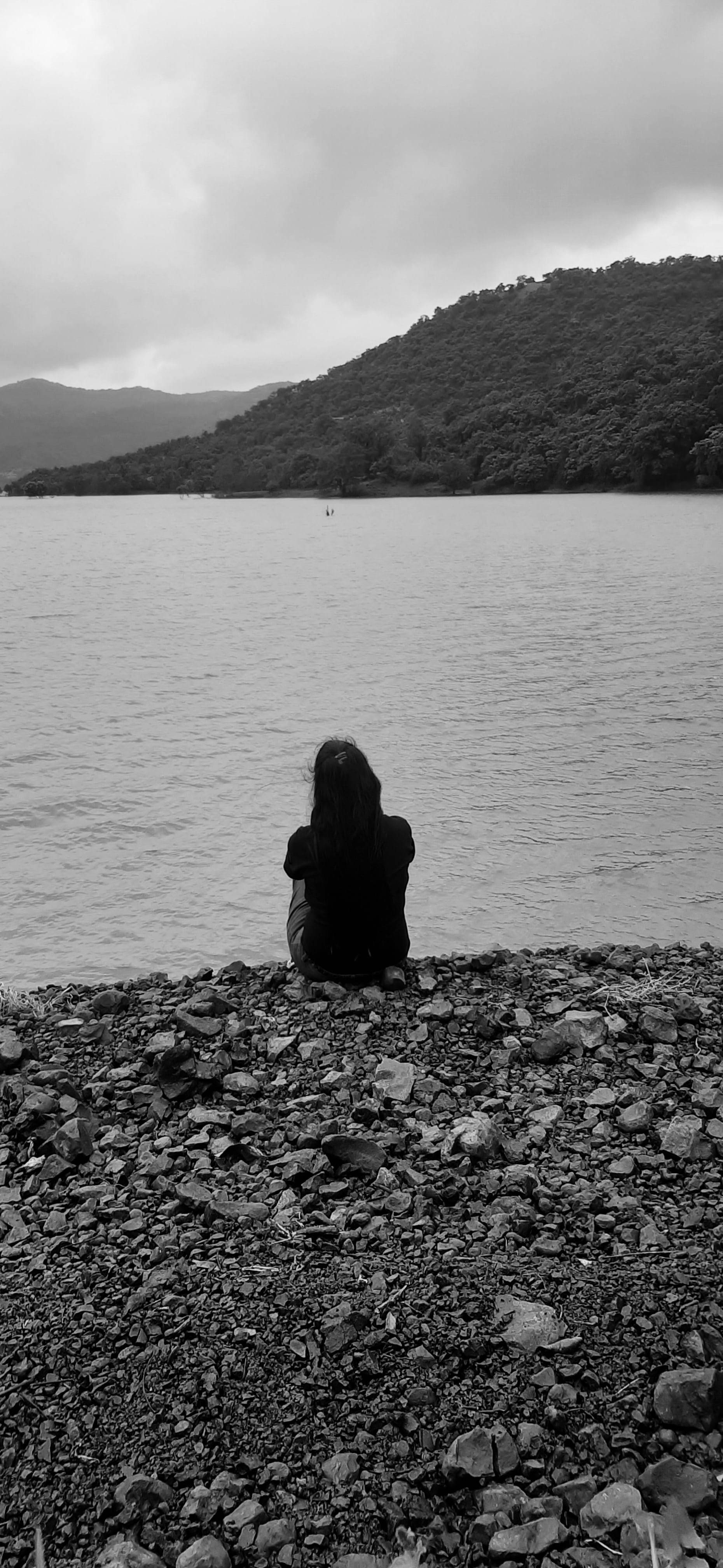 Depressed 桌布 壁紙 — 一個孤獨的身影坐在岩石海岸上，凝視著平靜的湖泊，湖邊有森林覆蓋的山丘，天空多雲。
