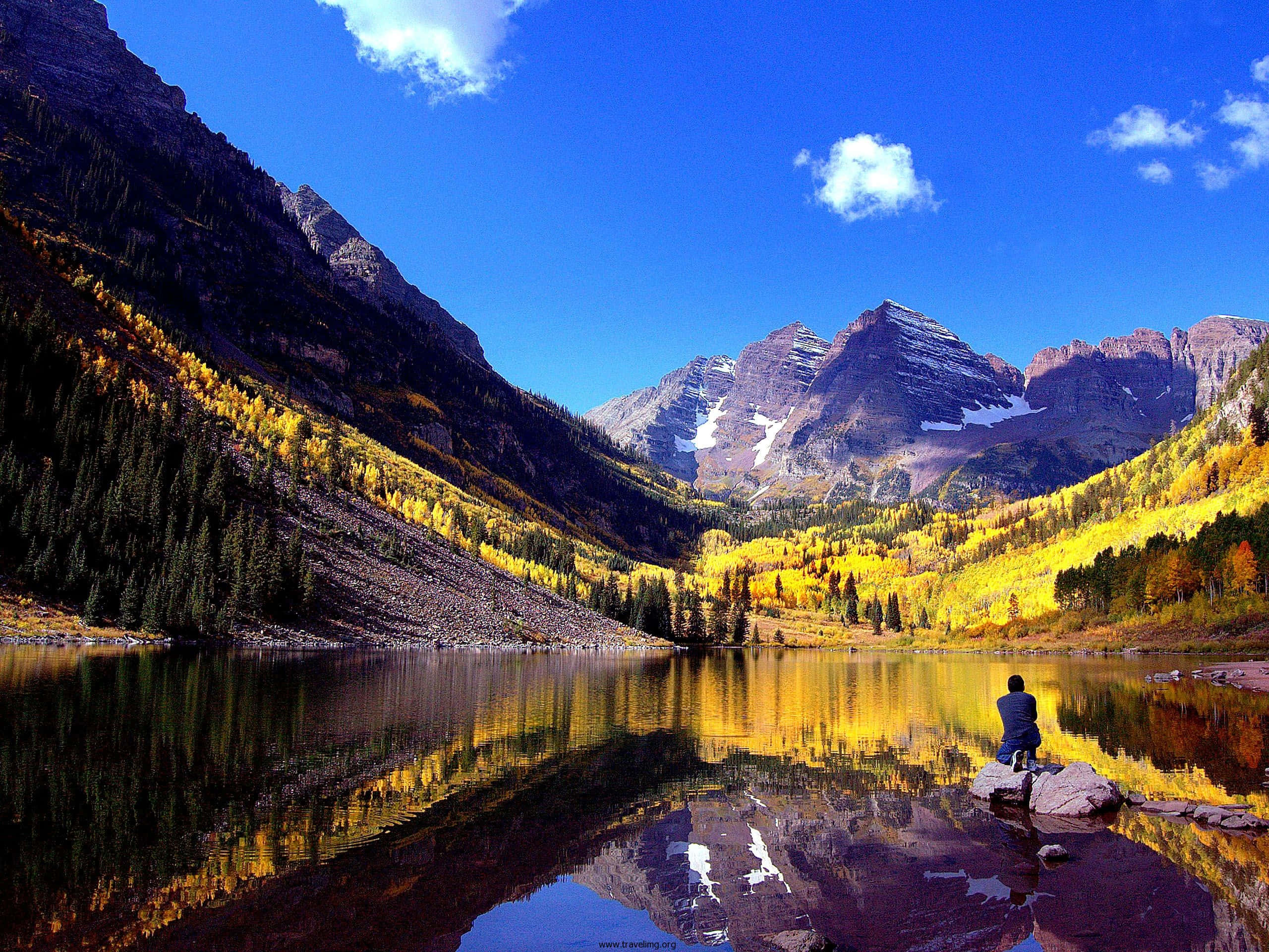 Aspen Colorado 桌布 壁紙 — 一個孤獨的身影坐在平靜的湖邊岩石上，湖水映照著金色的秋樹和白雪覆蓋的山脈，天空清澈湛藍。