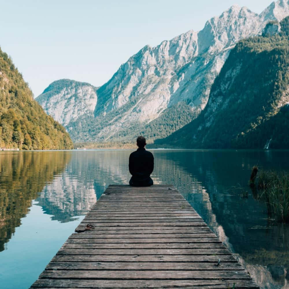 Alone 桌布 壁紙 — 一個孤獨的身影坐在木碼頭盡頭，靜靜沉思，俯瞰著寧靜的山湖，湖面倒映著高聳的山峰。