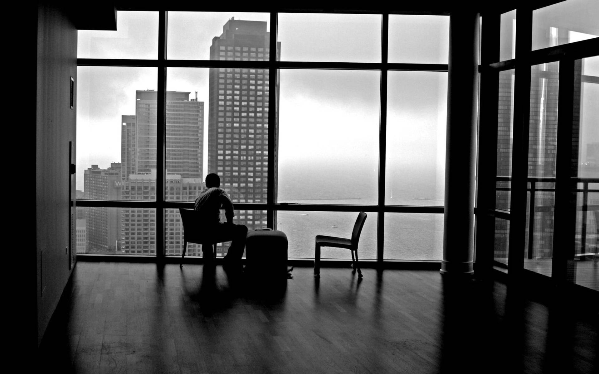 Alone 桌布 壁紙 — 一個孤獨的身影坐在椅子上，背對廣闊的窗戶，窗外是城市天際線和水面，天空多雲。