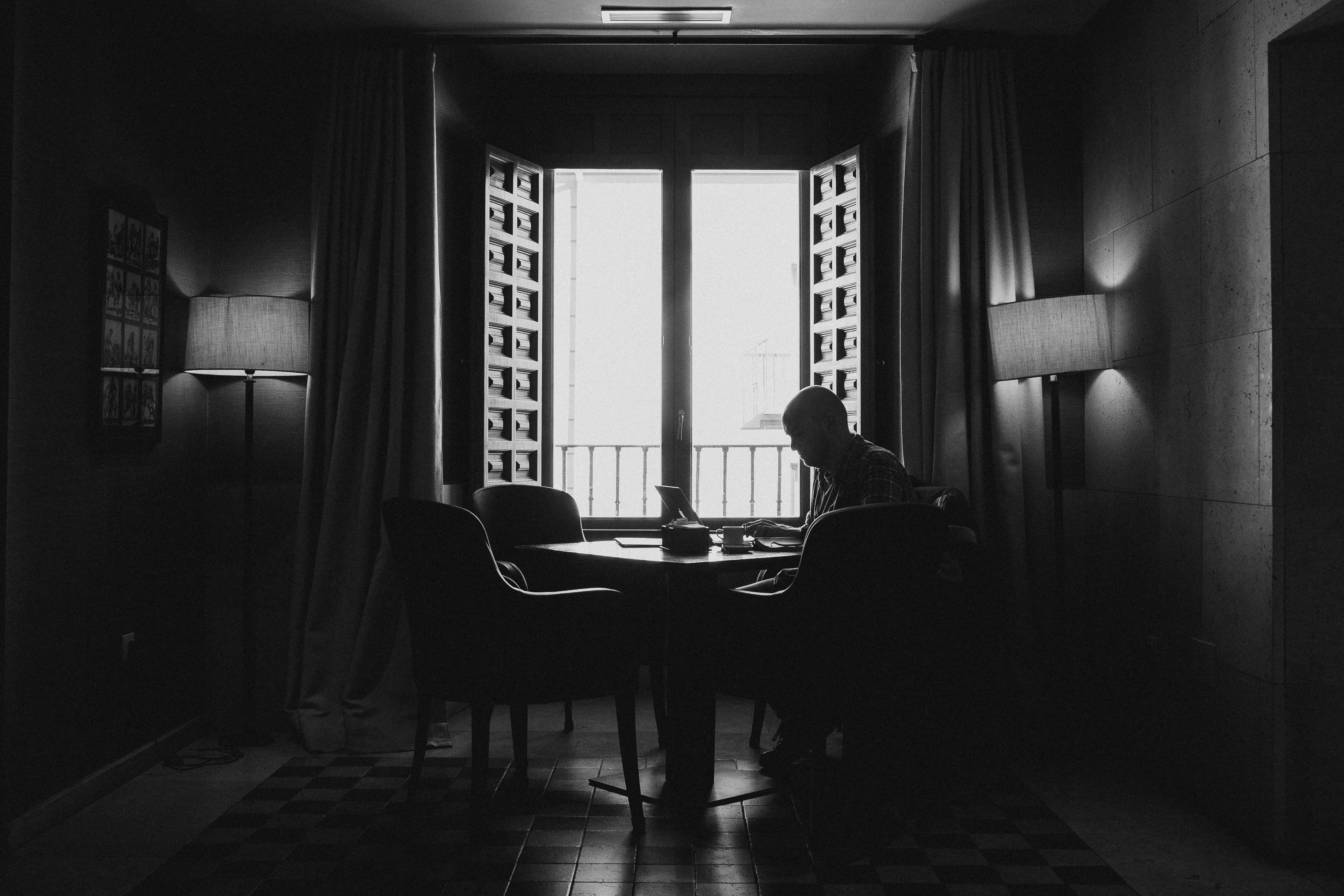 Aesthetic Black And White 桌布 壁紙 — 一個孤獨的身影坐在燈光昏暗的房間裡的桌子旁，在明亮的窗光映襯下形成剪影。