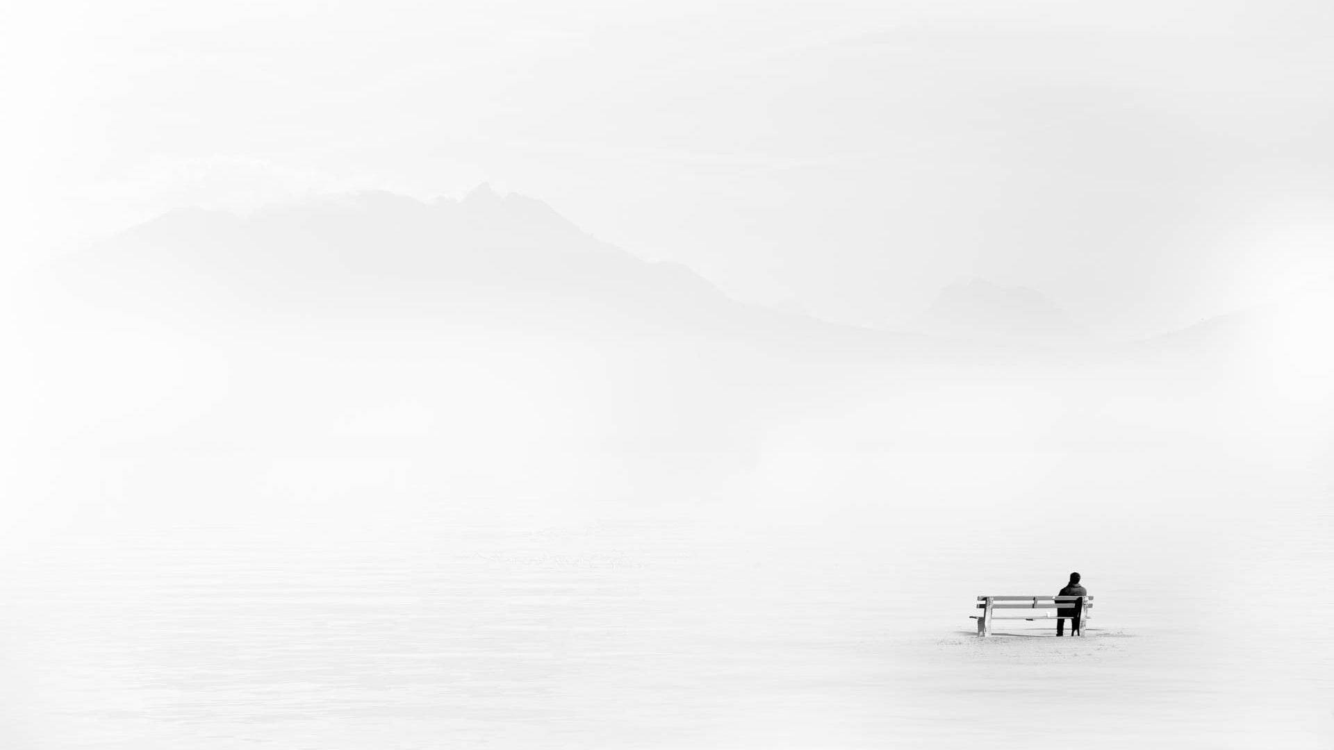 Sitting Alone 桌布 壁紙 — 一個孤獨的身影坐在長椅上，眼前是廣闊的雪景，遠處有霧氣繚繞的山巒。