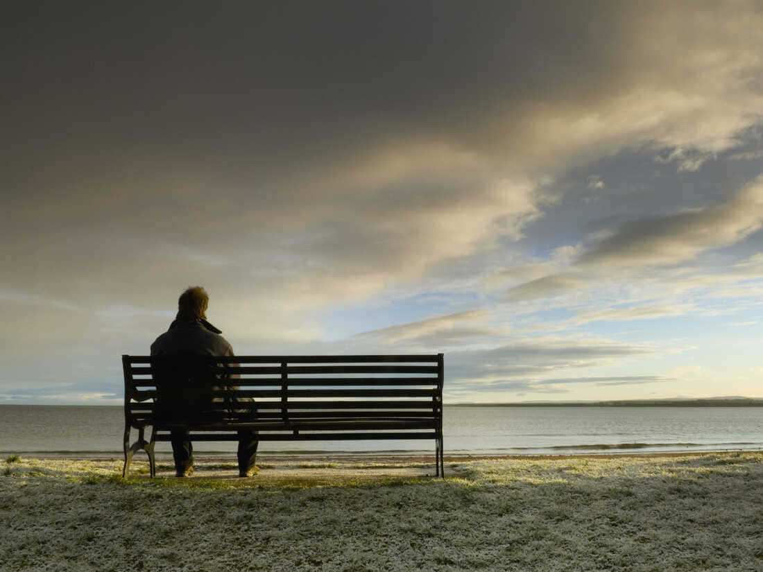 Lonely 桌布 壁紙 — 一個孤獨的身影坐在長椅上，面向廣闊的水域，背景是黃昏時分戲劇性的多雲天空。