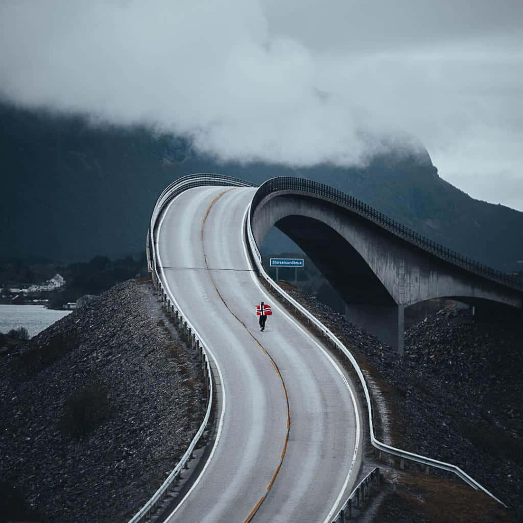 Road 桌布 壁紙 — 一個孤獨的身影拿著挪威國旗，在陰雲密布的天空下沿著蜿蜒的山路行走。