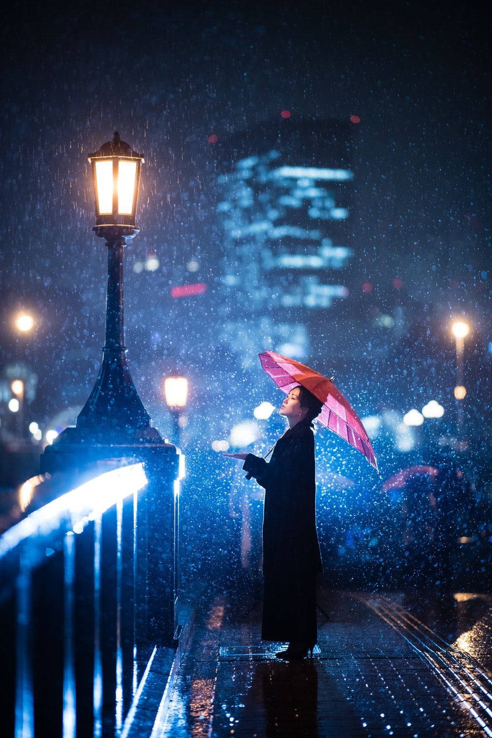 Screen 桌布 壁紙 — 一個孤獨的身影拿著粉紅色雨傘，在夜晚的城市橋樑上，站在發光路燈下的大雨中。