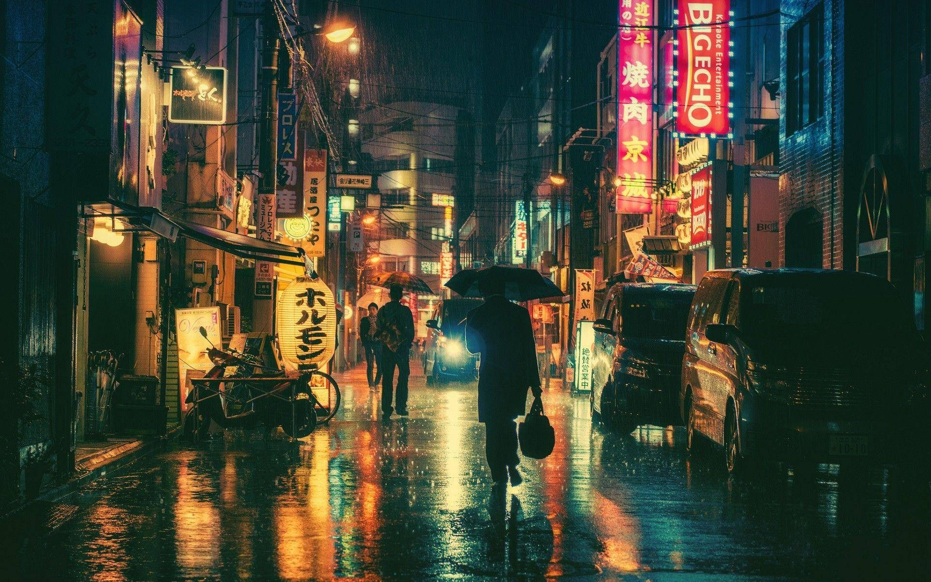 日本 桌布 壁紙 — 一個孤獨的身影撐著傘，在夜晚霓虹燈照亮、雨水浸濕的東京小巷中行走，映照出鮮豔的招牌。
