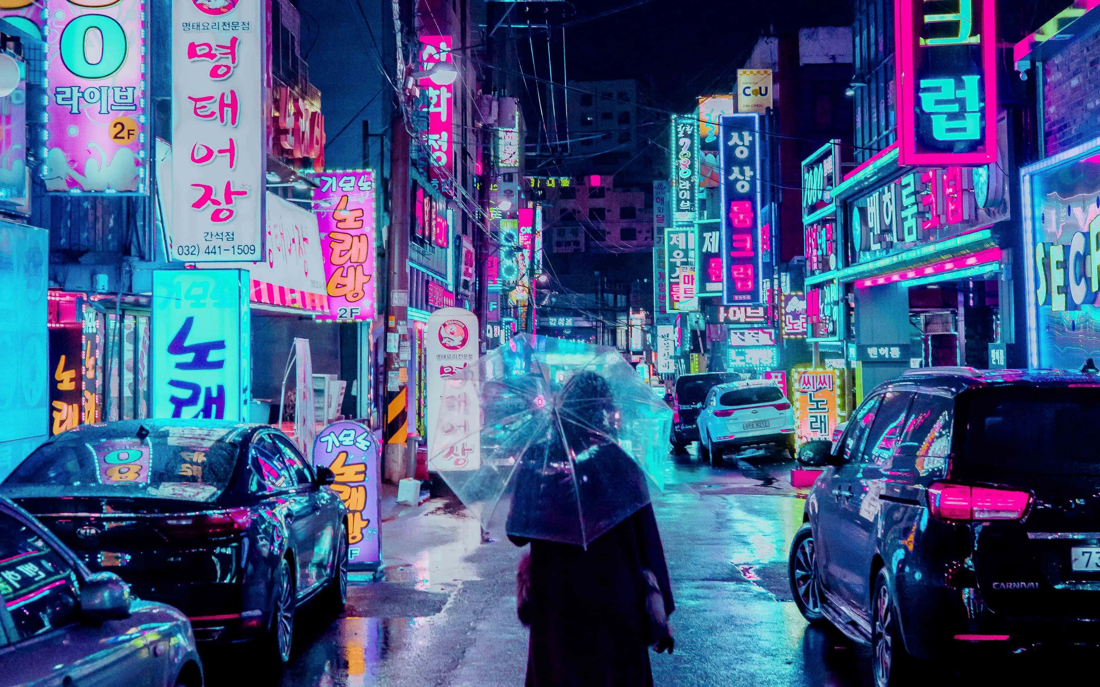 美學 桌布 壁紙 — 一個孤獨的身影撐著傘，夜晚走在霓虹燈閃爍的韓國街道上，潮濕的路面反射出鮮豔的粉紅、藍色和綠色燈光。