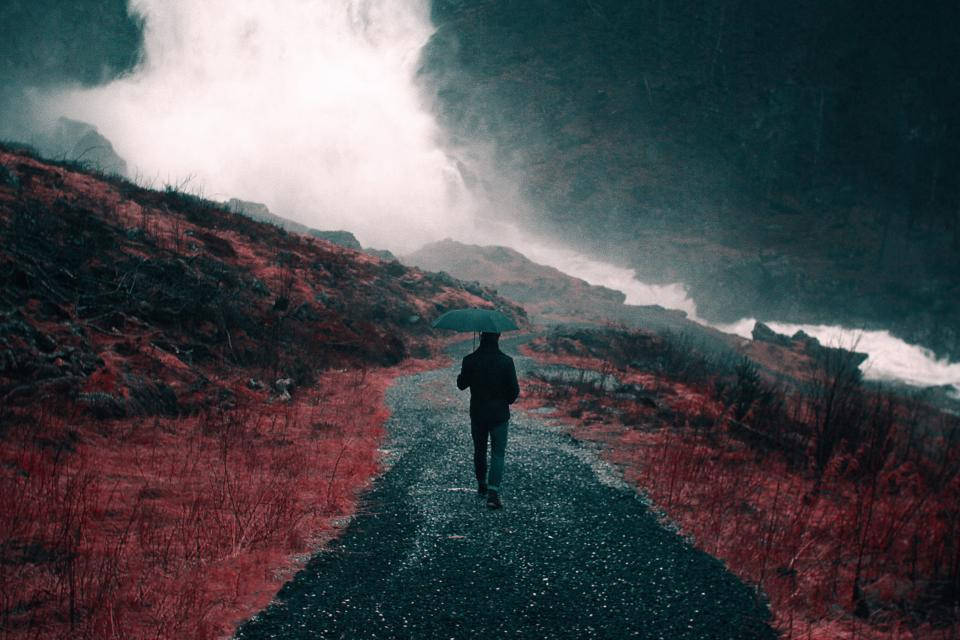 Sad Alone 桌布 壁紙 — 一個孤獨的身影撐著傘，走在一條濕漉漉的小徑上，穿過霧氣瀰漫的火山地貌，紅土與烏雲交織。