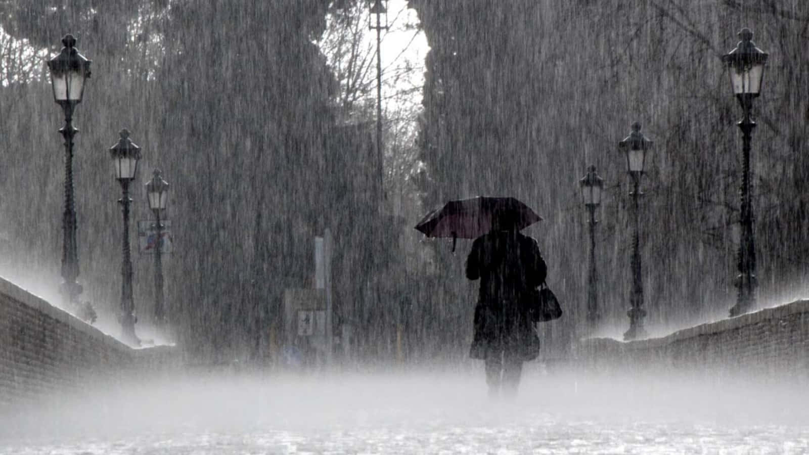 Raining 桌布 壁紙 — 一個孤獨的身影撐著深色雨傘，在霧氣瀰漫的都市街道上冒著傾盆大雨行走，街道兩旁是復古路燈。