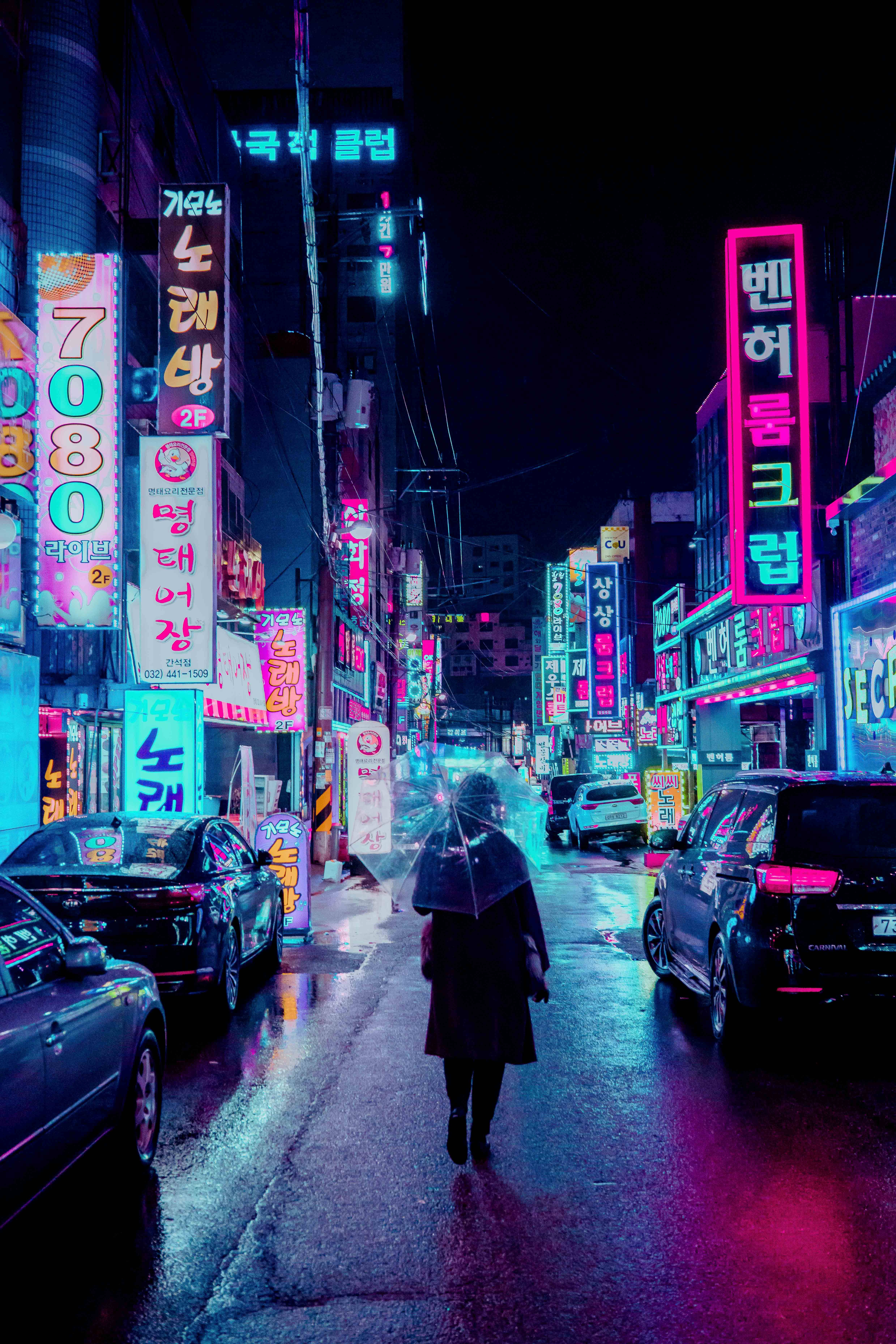 城市 桌布 壁紙 — 一個孤獨的身影撐著雨傘，在夜晚沿著被雨水浸濕、霓虹燈閃爍的小巷行走，周圍是發光的韓文招牌和停泊的汽車。