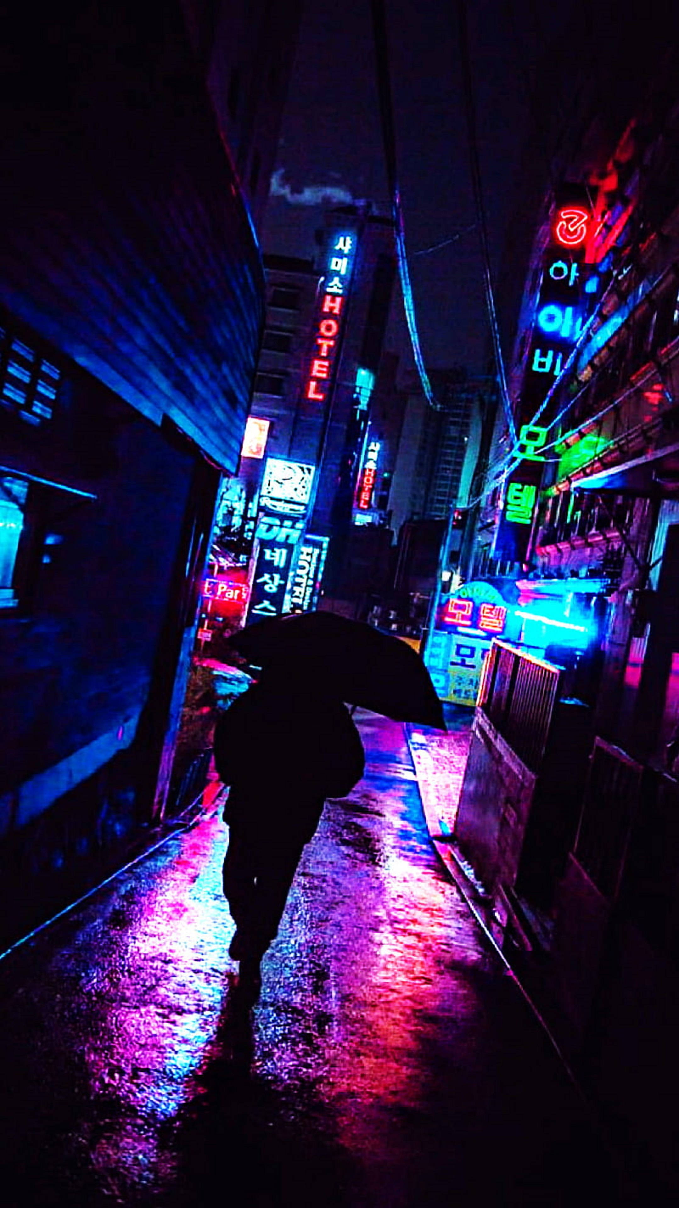 4K 霓虹 iPhone 桌布 壁紙 — 一個孤獨的身影撐著雨傘，在夜晚走過一條被霓虹燈浸透、雨水浸濕的小巷，反射出鮮豔的粉紅和藍色燈光。