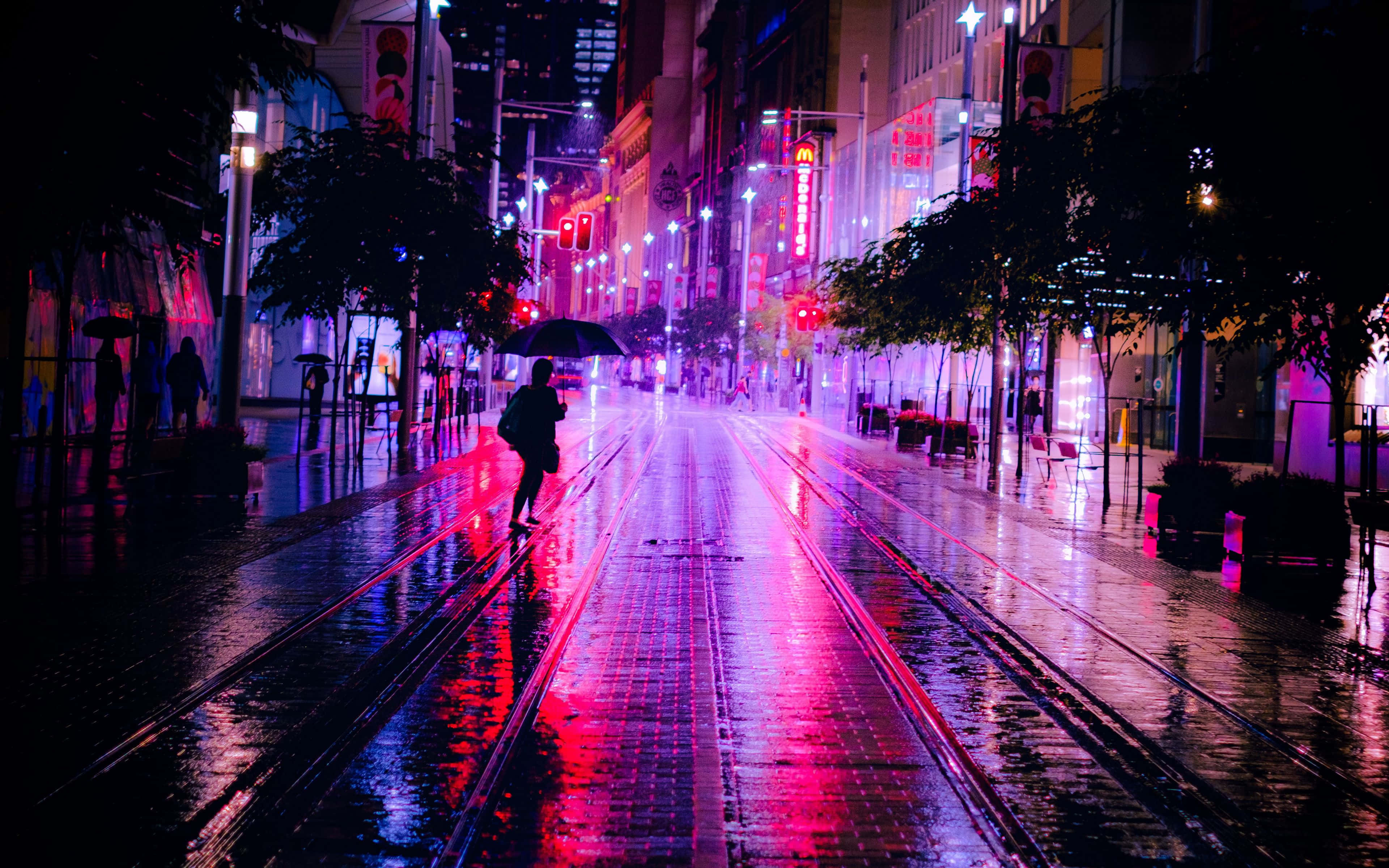 Night Street 桌布 壁紙 — 一個孤獨的身影撐著雨傘，在夜晚霓虹燈閃爍、雨水浸濕的城市街道上行走，反射出鮮豔的粉紅色和藍色燈光。