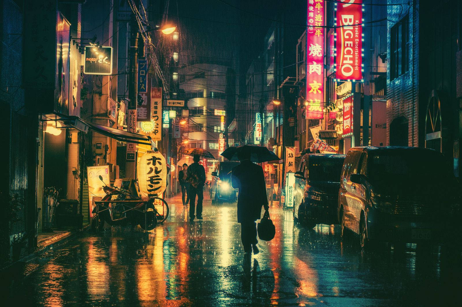 東京 桌布 壁紙 — 一個孤獨的身影撐著雨傘，在夜晚霓虹燈照亮、雨水濕滑的日本街道上行走，映照出鮮豔的招牌。