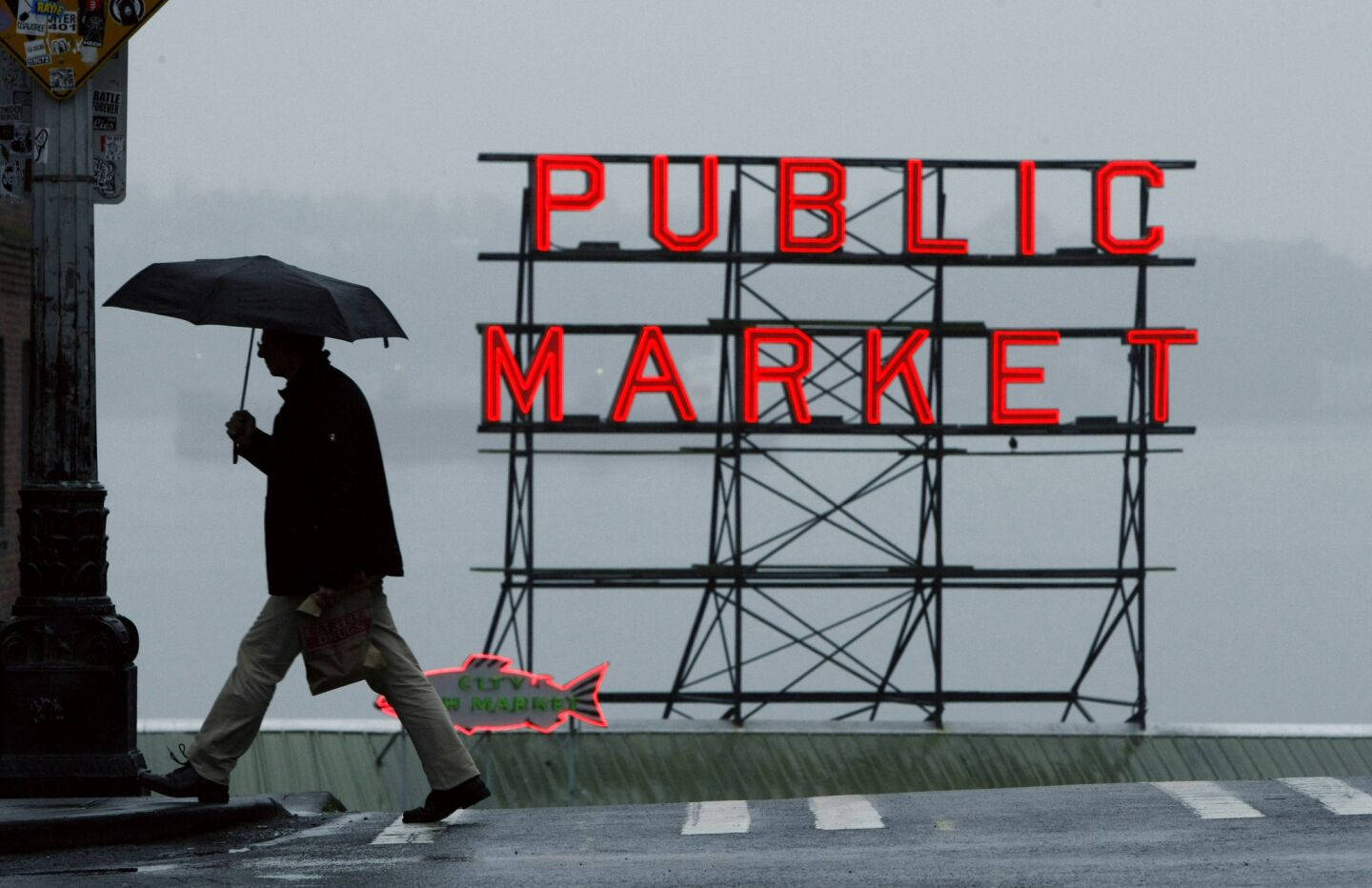 Pike Place Market 桌布 壁紙 — 一個孤獨的身影撐著雨傘，在雨天的城市街道上走過一個發光的紅色「PUBLIC MARKET」標誌。