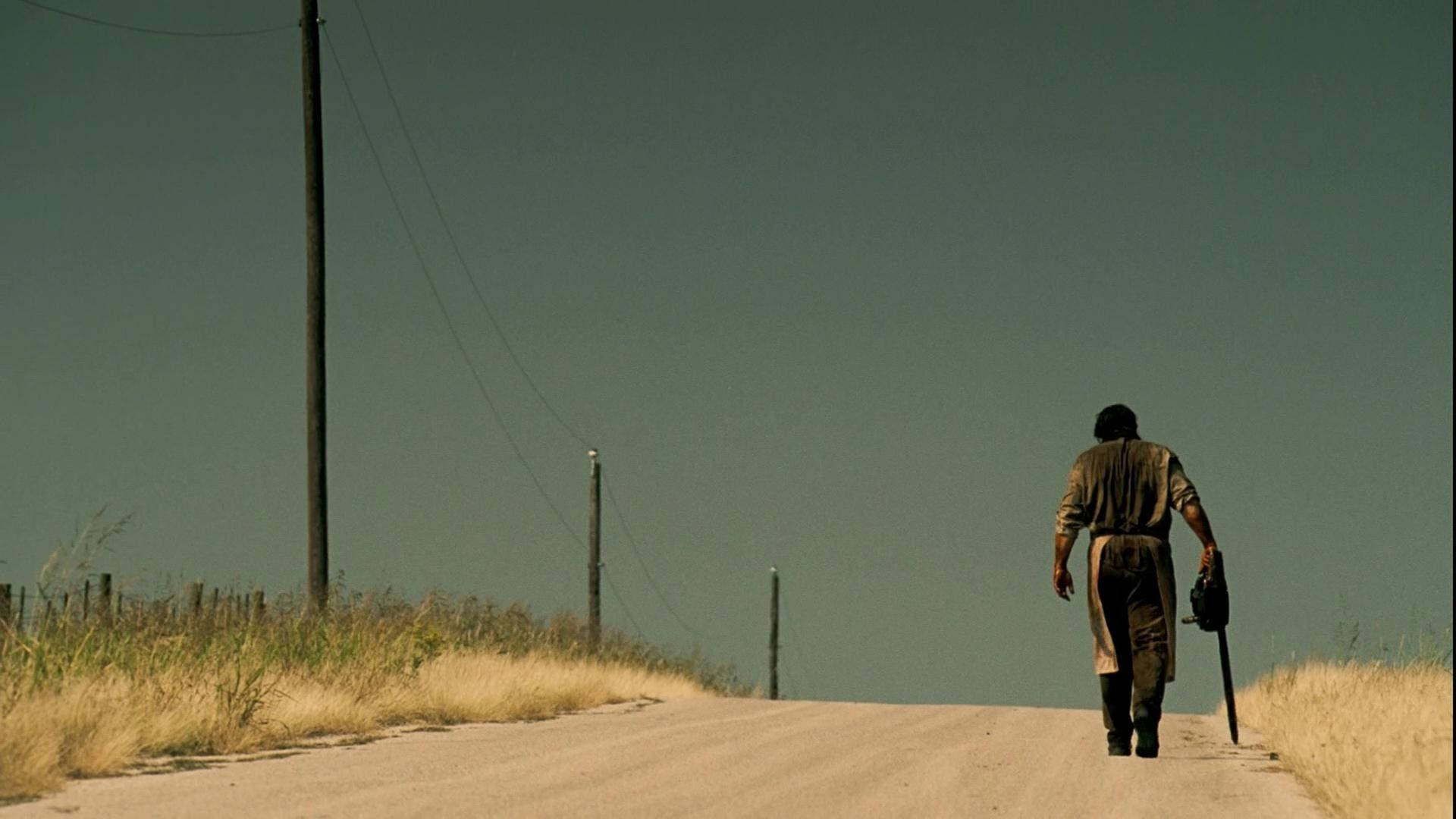 Leatherface 桌布 壁紙 — 一個孤獨的身影沿著塵土飛揚的鄉村道路走遠，頭頂是廣闊而柔和的天空。