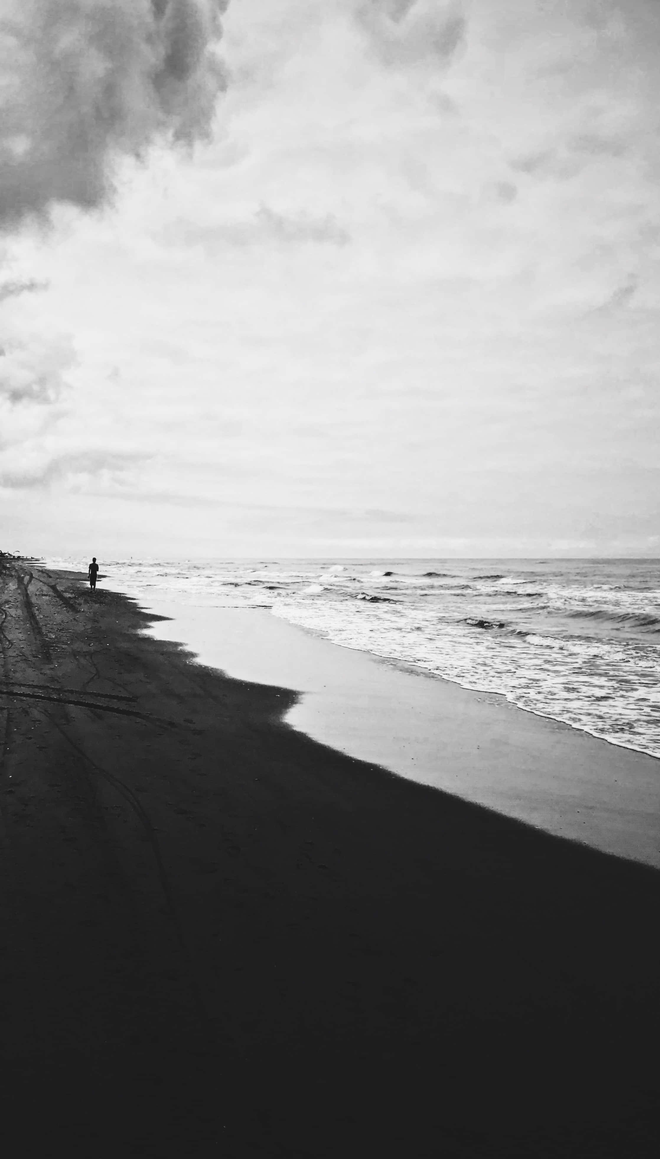 Black And White Beach 桌布 壁紙 — 一個孤獨的身影沿著黑暗的沙灘行走，溫柔的海浪在陰天中翻滾。