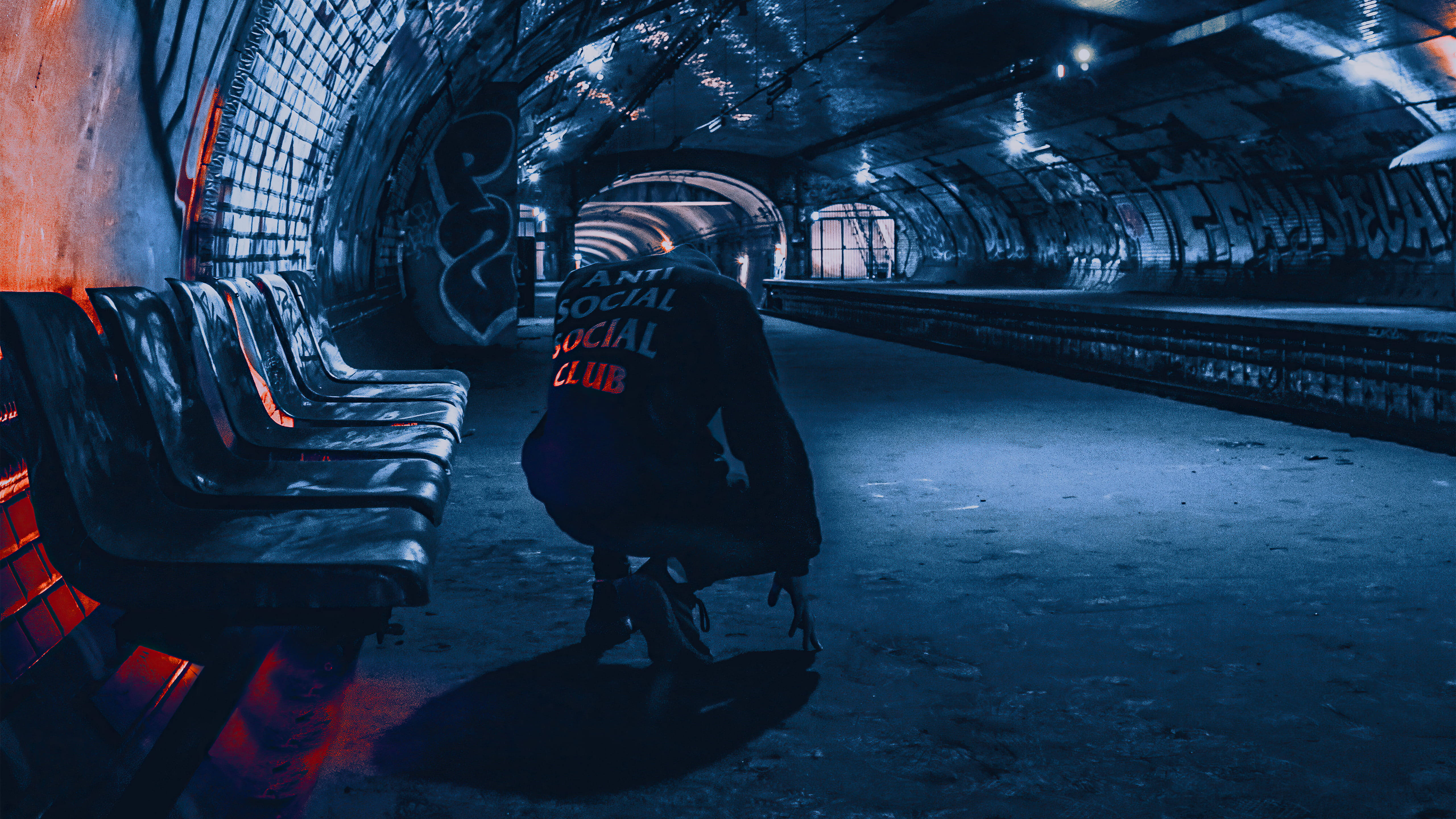 Social 桌布 壁紙 — 一個孤獨的身影穿著帶有紅色文字的黑色外套，蹲在一個被塗鴉覆蓋的地鐵隧道中，被冷藍色和橙色燈光照亮。