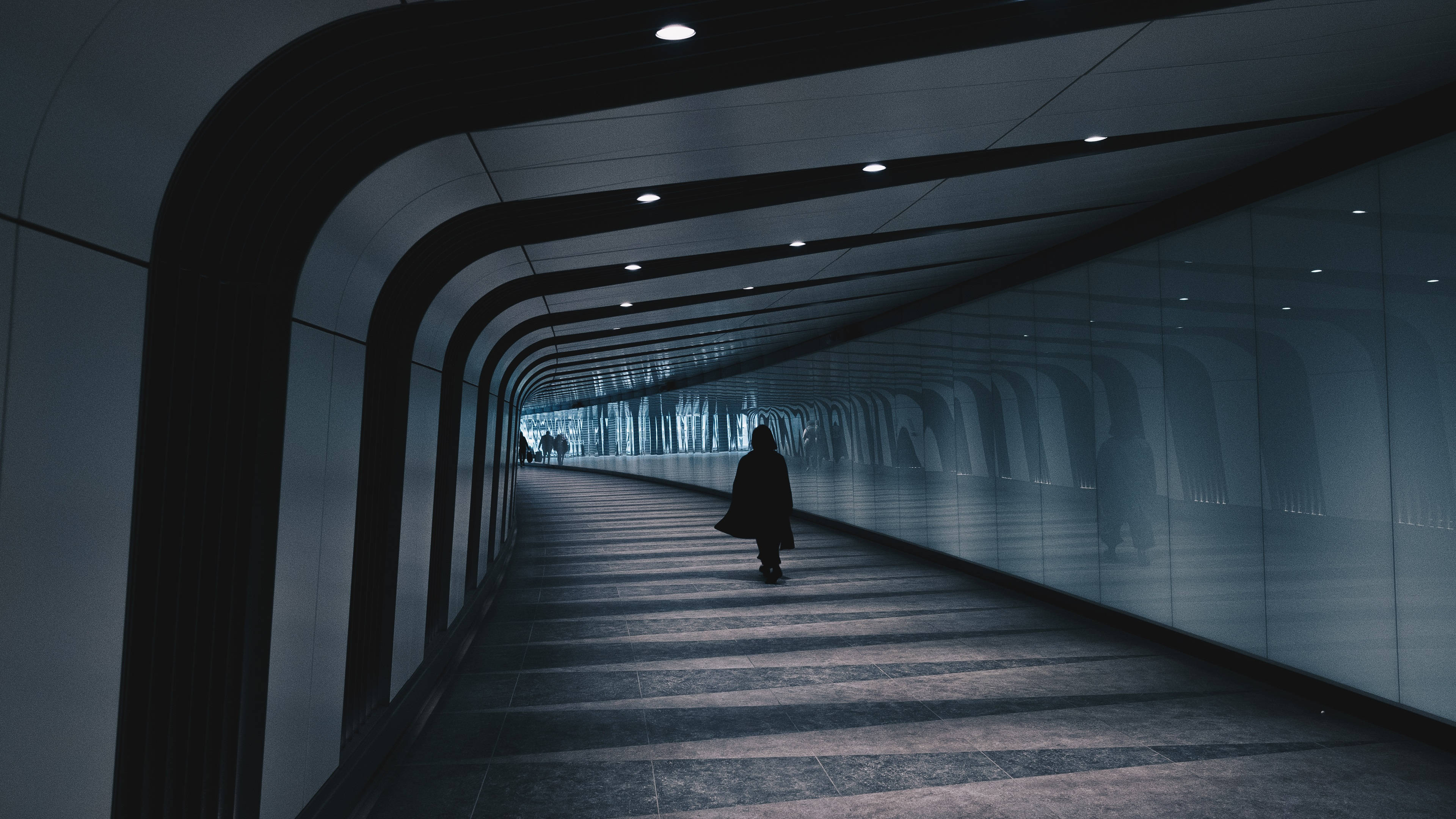 tunnel 桌布 壁紙 — 一個孤獨的身影穿著深色外套，沿著一條由天花板燈照亮的長彎曲玻璃牆隧道行走。