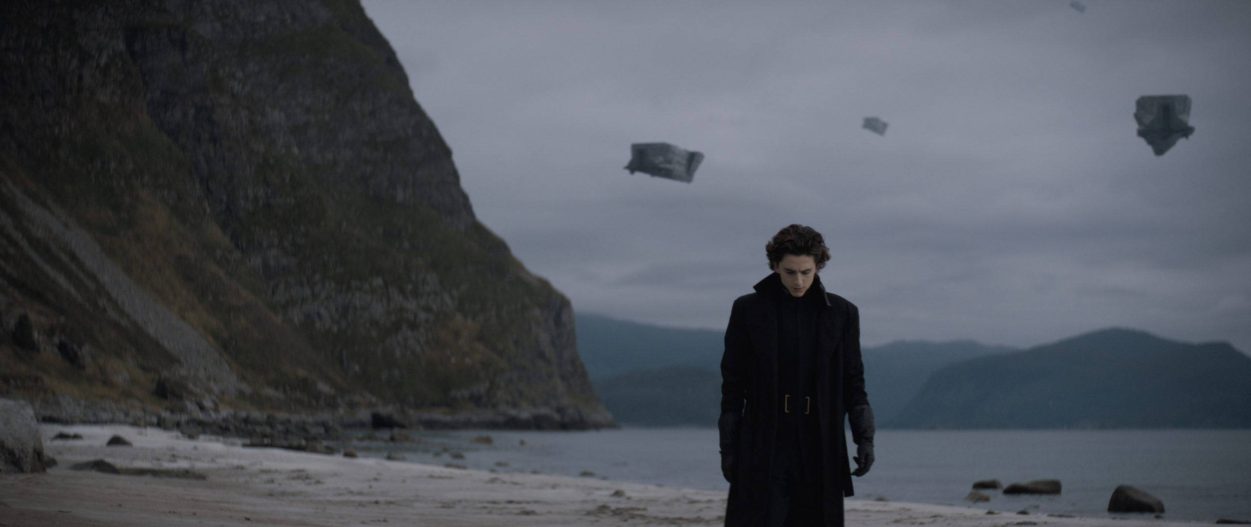 Dune Movie 桌布 壁紙 — 一個孤獨的身影穿著深色外套，站在霧氣瀰漫的海岸上，浮石般的物體在崎嶇的懸崖和平靜的海面上飄浮。