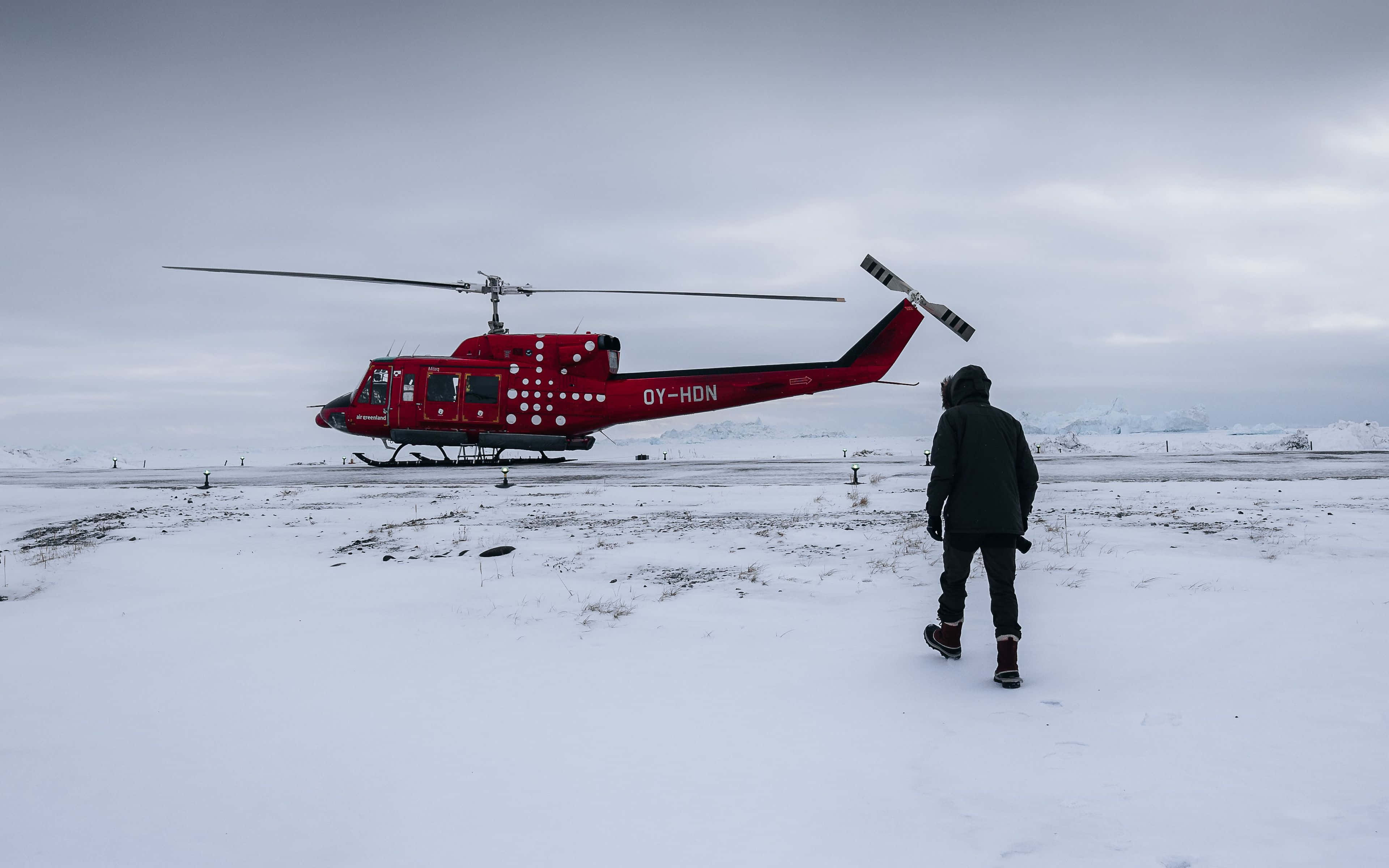 4K Helicopters 桌布 壁紙 — 一個孤獨的身影穿著深色裝備，在陰天之下，朝着一架停泊在廣闊雪原上的鮮紅色直升機走去。