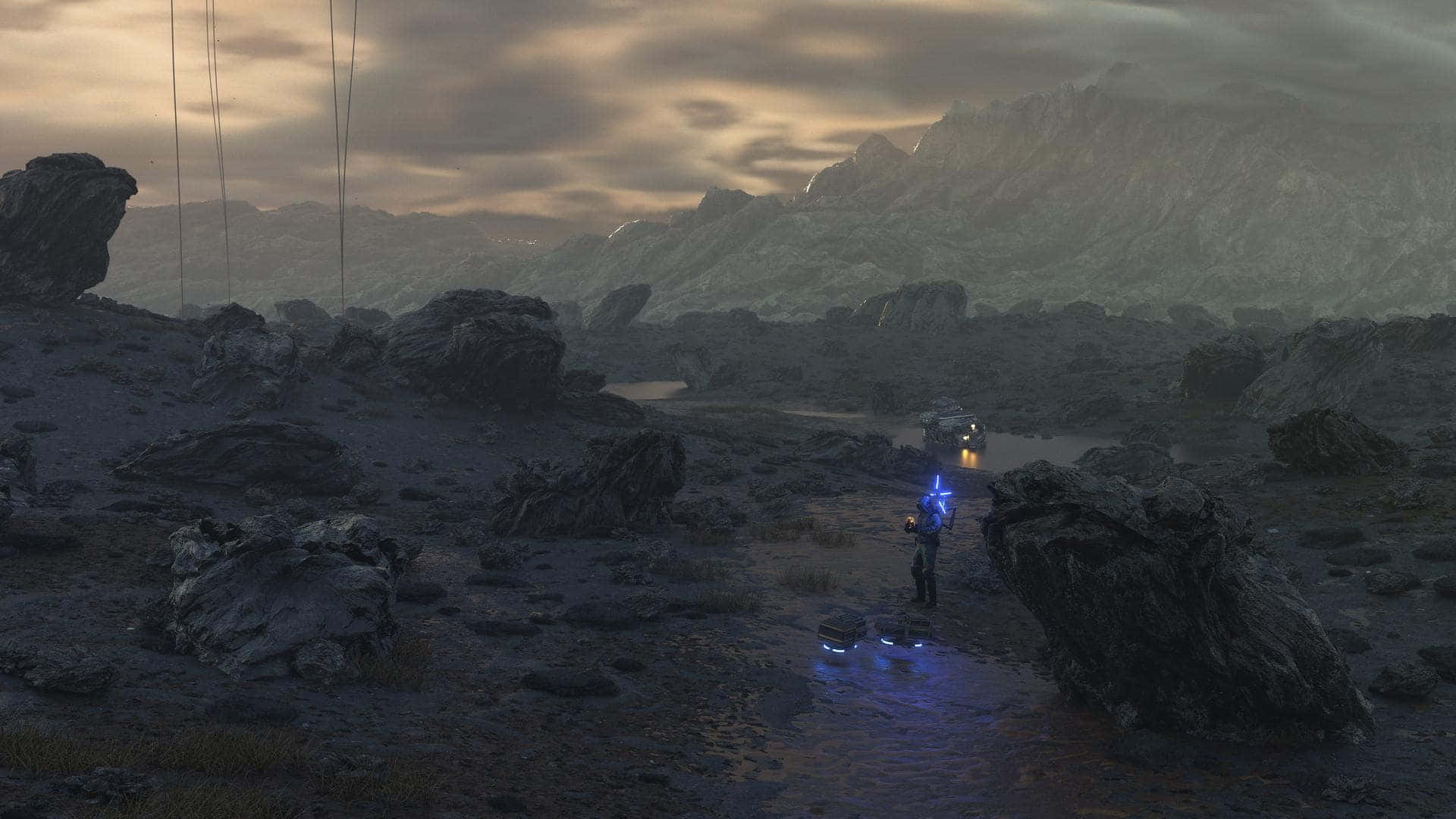 Death Stranding Pc 桌布 壁紙 — 一個孤獨的身影穿著發光的藍色盔甲，站在多雲天空下荒涼的岩石景觀中，遠處有一輛車正在靠近。
