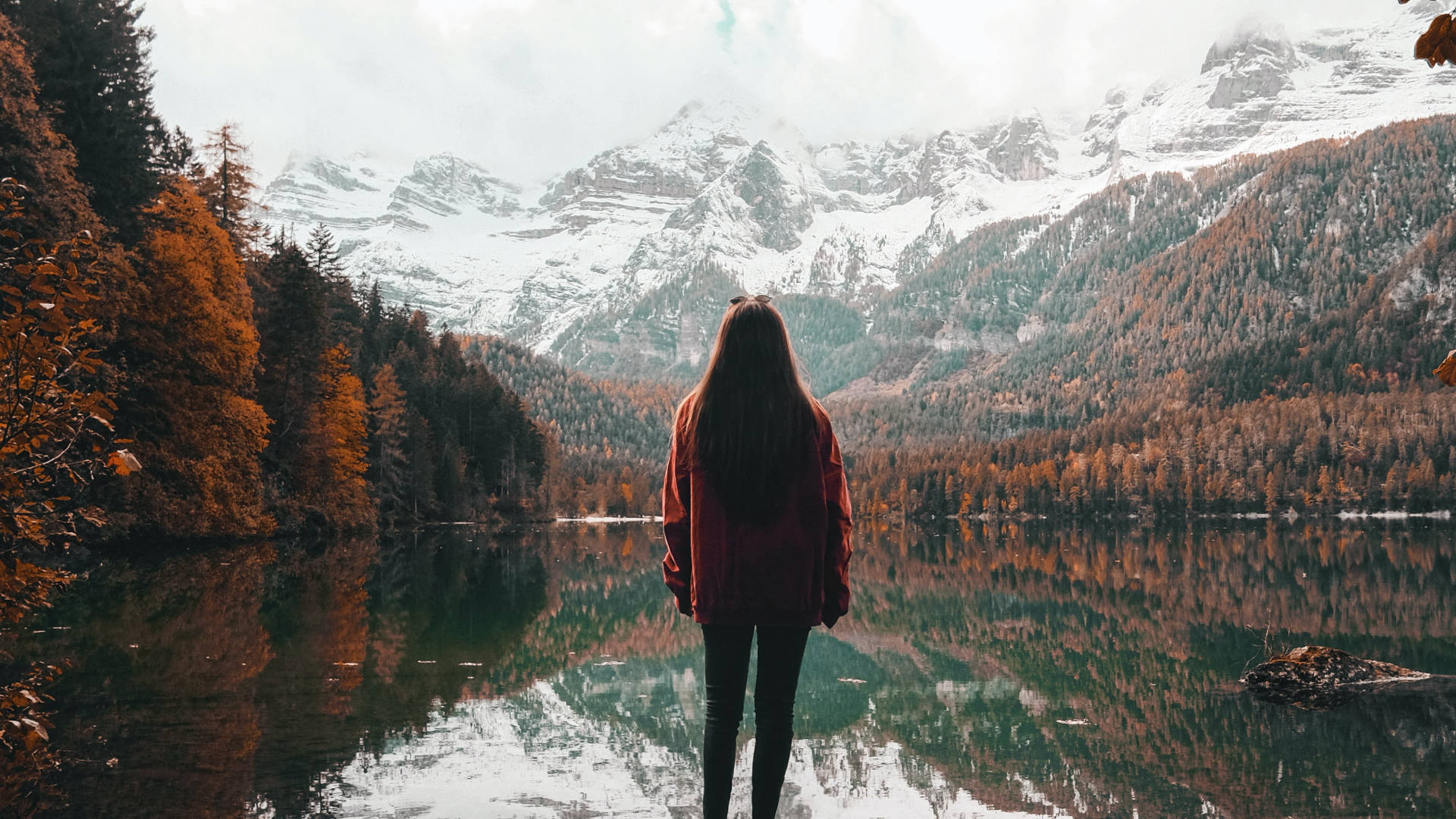 Lake 桌布 壁紙 — 一個孤獨的身影穿著紅色外套，站在平靜的湖邊，湖面倒映著秋天的樹木和積雪的山峰，天空多雲。