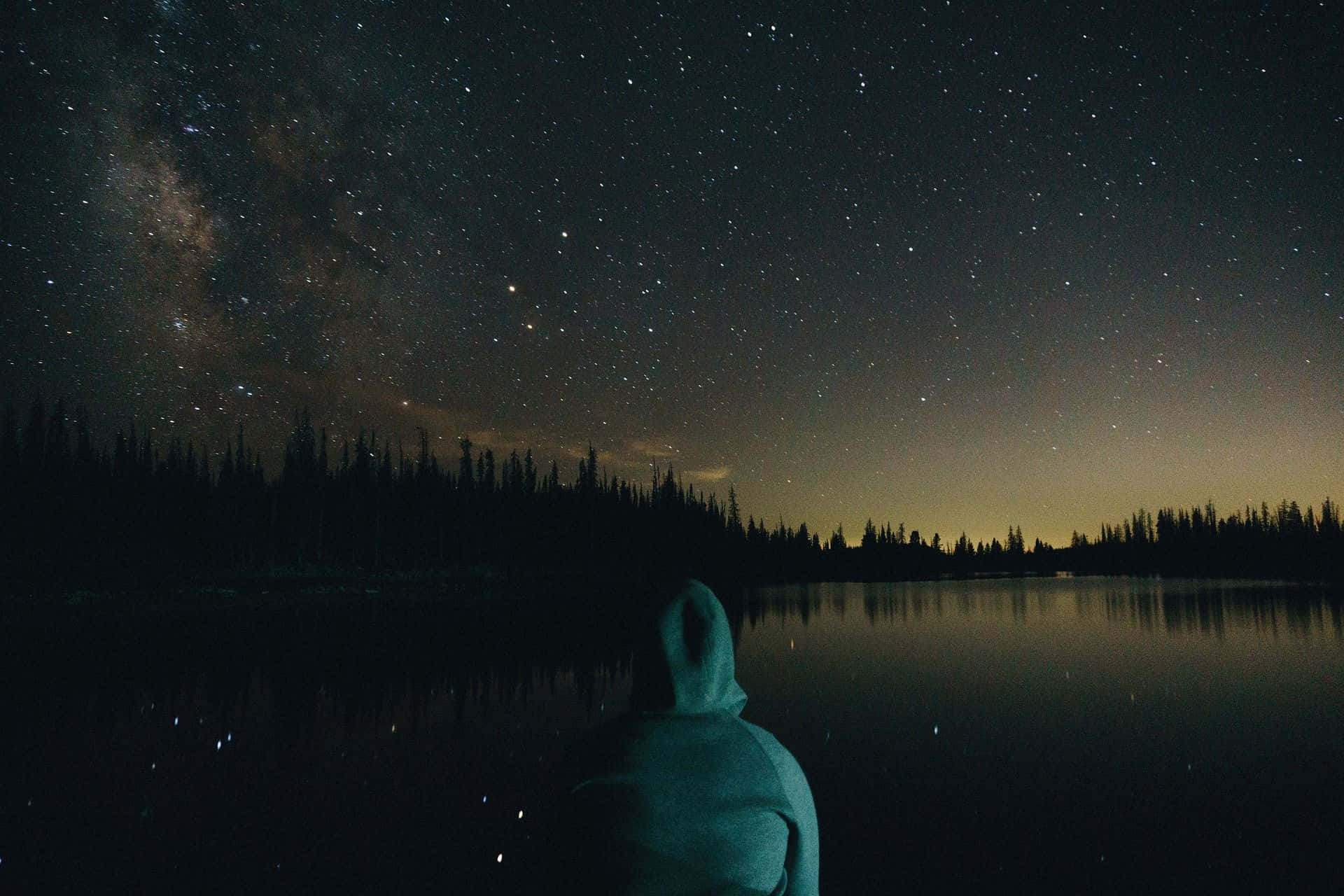 Lonely 桌布 壁紙 — 一個孤獨的身影穿著連帽衫，凝視著星空下寧靜湖面中反射的銀河。