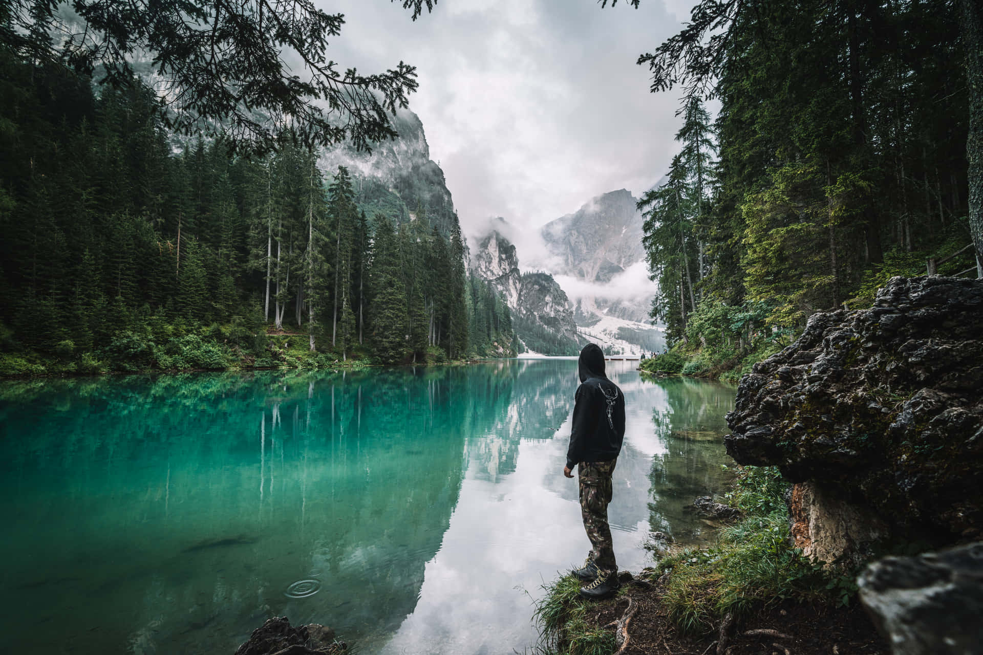 穿著連帽衫的人站在碧綠的湖邊，背景是森林和山巒。 — Google Meet 壁紙
