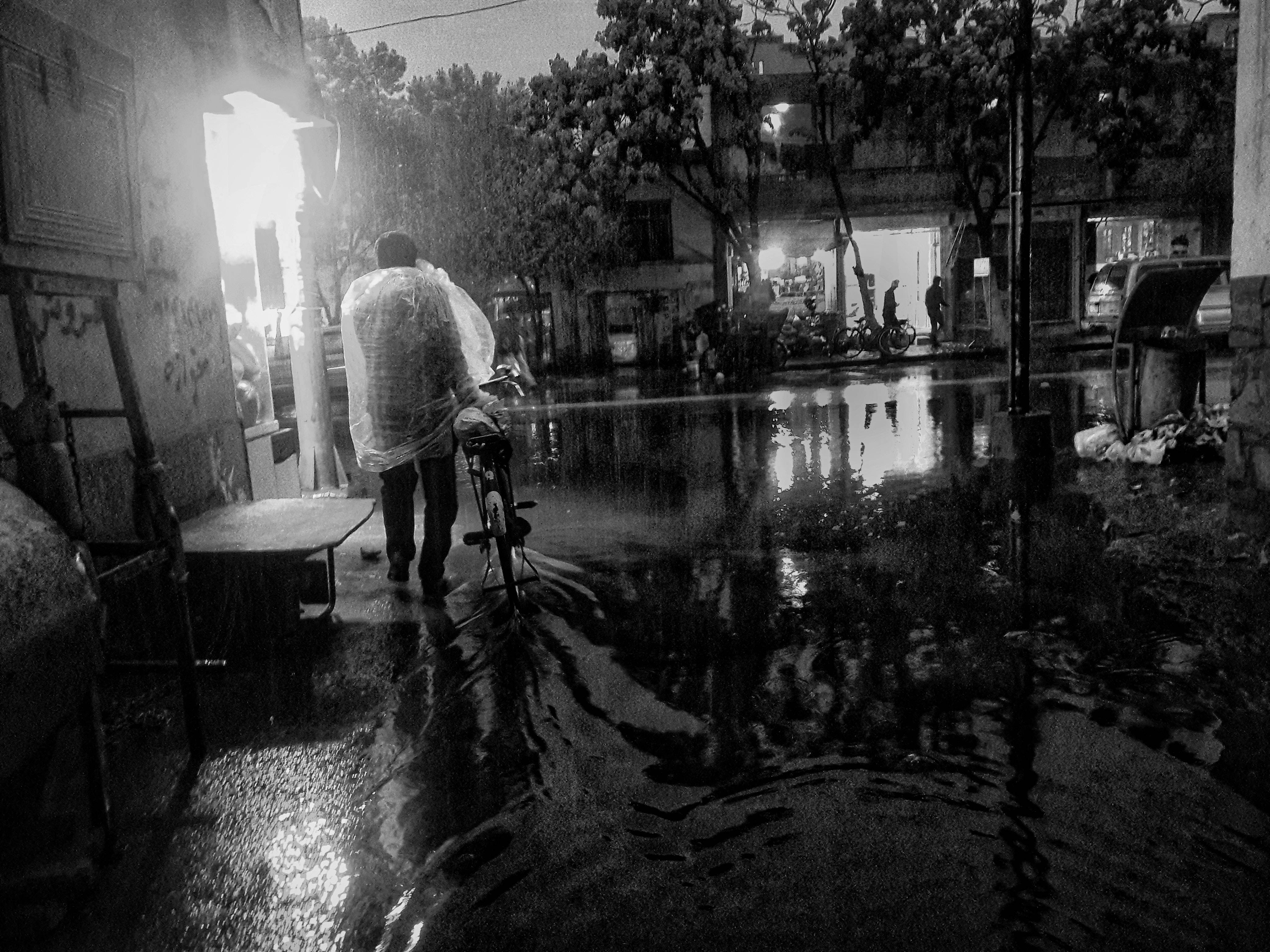 Kabul 桌布 壁紙 — 一個孤獨的身影穿著雨衣，在夜晚推著腳踏車穿過被水淹沒的城市街道，濕漉漉的路面上反射著閃爍的光芒。