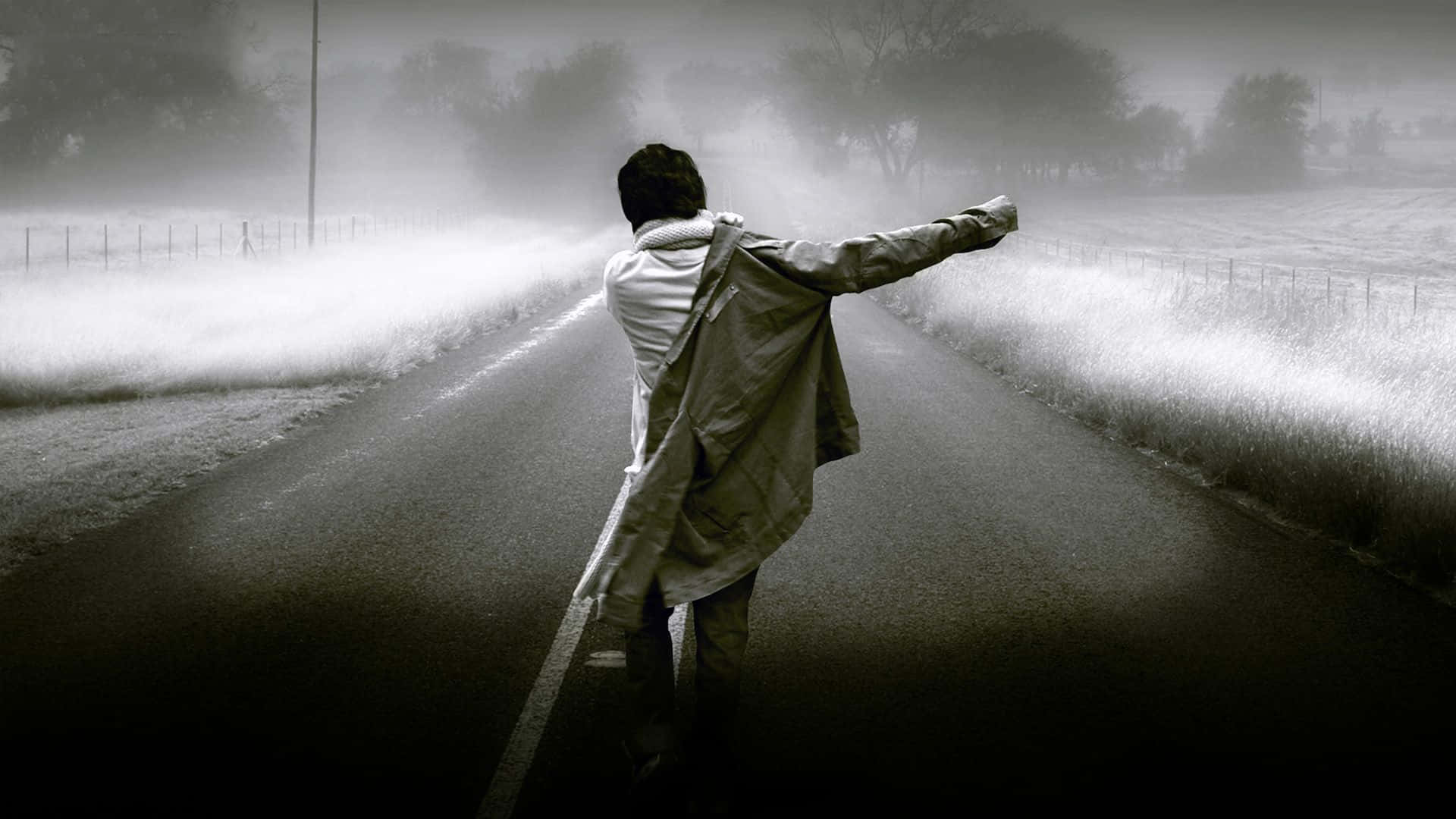 Alone 桌布 壁紙 — 一個孤獨的身影穿著風衣，站在霧氣瀰漫的鄉村道路上，雙臂伸展，周圍是霧濛濛的田野和光禿禿的樹木。