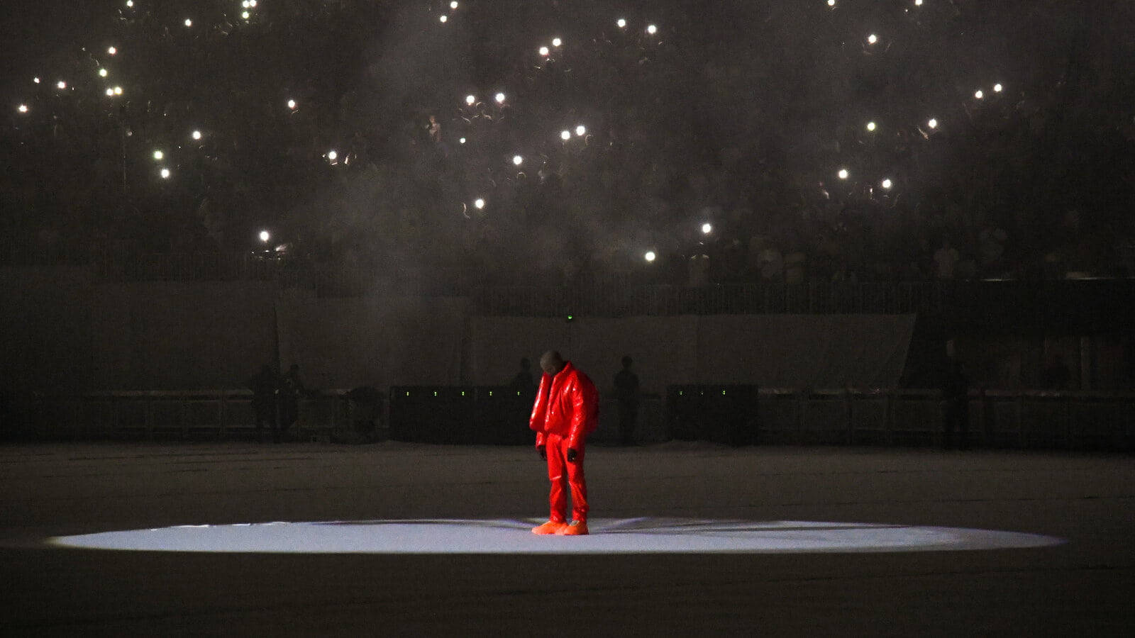 Kanye West Ye 桌布 壁紙 — 一個孤獨的身影穿著鮮紅色西裝，站在舞台中央的聚光燈下，黑暗的場館上方有數千支手機燈光閃爍。