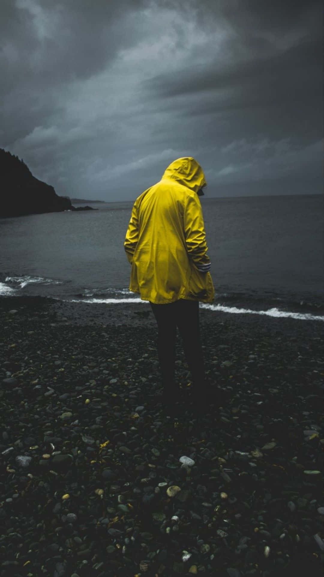 Yellow Raincoat 桌布 壁紙 — 一個孤獨的身影穿著鮮黃色雨衣，站在深色卵石海灘上，面對著厚重烏雲下的暴風雨大海。