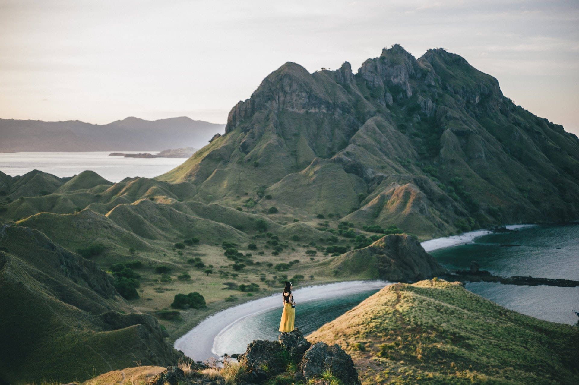 Mountain Landscape 桌布 壁紙 — 一個孤獨的身影穿著黃色連衣裙，站在長滿草的山丘上，俯瞰著新月形海灘和鋸齒狀的綠色山脈，正值黃金時刻。