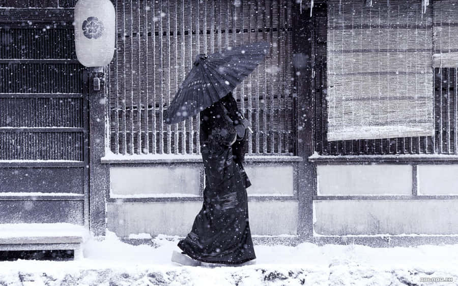 High Resolution Snow 桌布 壁紙 — 一個孤獨的身影穿著黑色和服，在飄雪中走過一座傳統日本建築，旁邊有竹簾和紙燈籠。