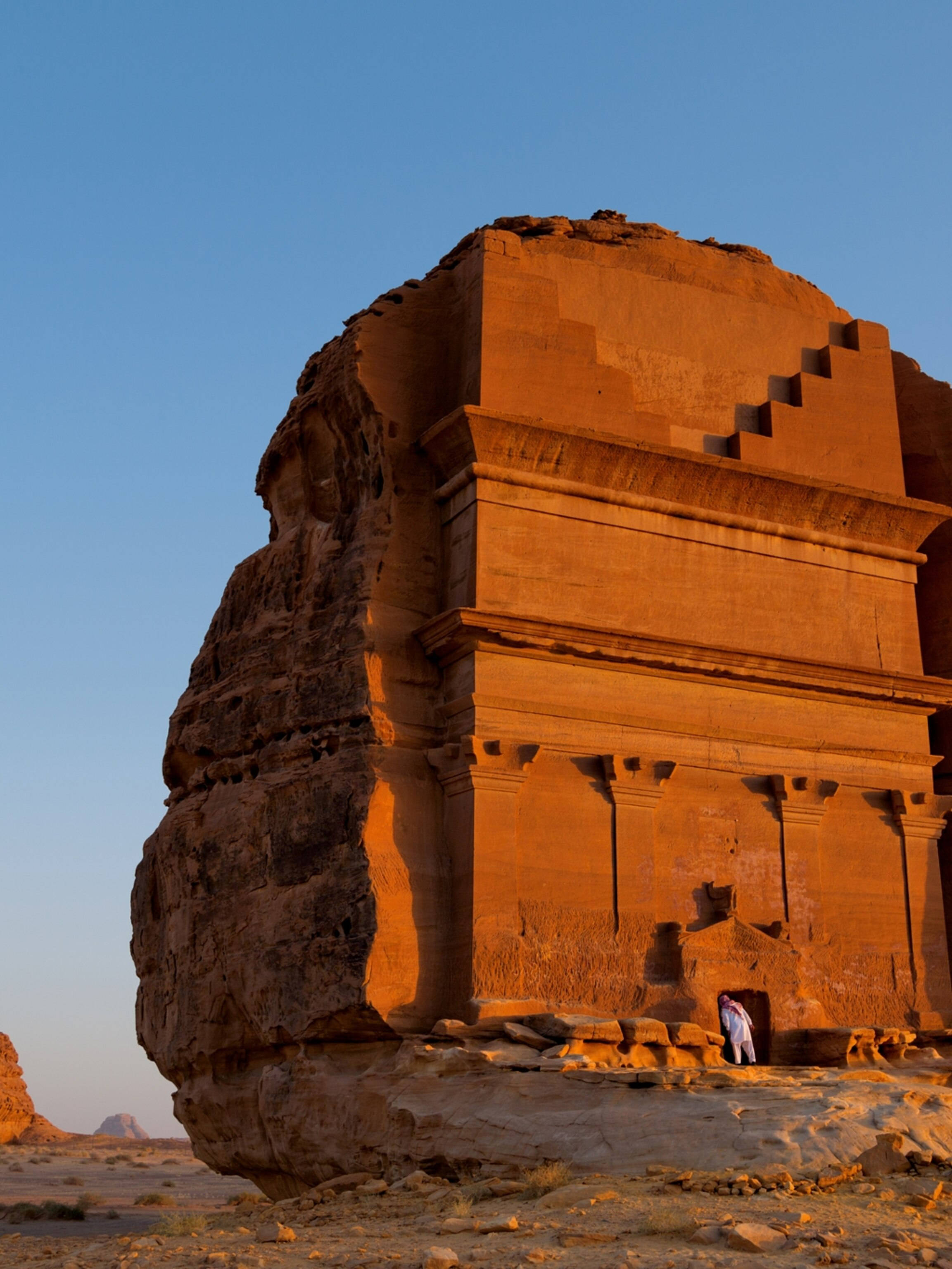 Saudi 桌布 壁紙 — 一個孤獨的身影站在一座古老的、陽光普照的砂岩寺廟前，該寺廟雕刻在沙漠懸崖上，背景是清澈的藍天。