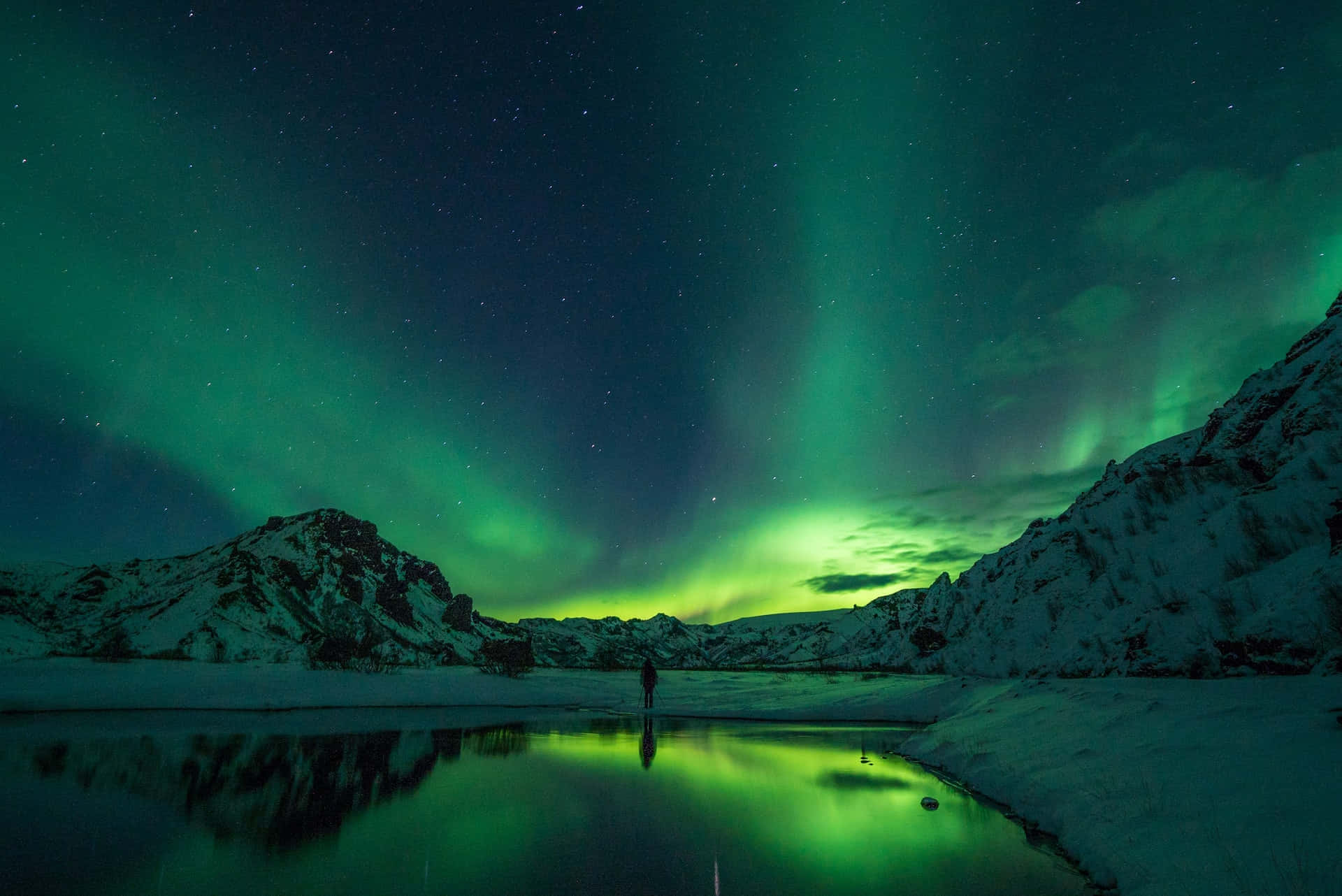 Iceland Desktop 桌布 壁紙 — 一個孤獨的身影站在冰凍的湖邊，腳下是燦爛的綠色北極光在繁星點點的冬日天空中舞動。