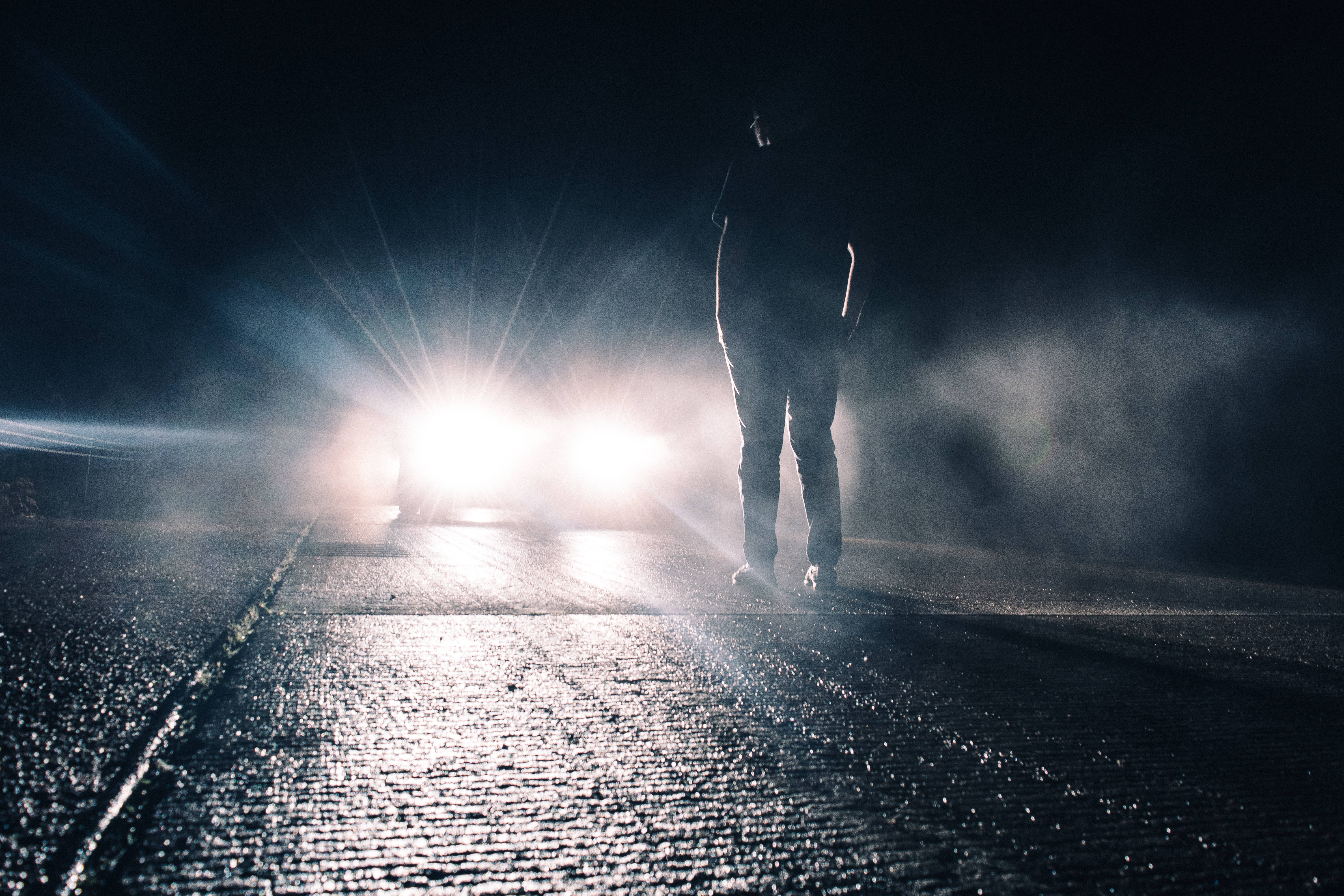 詭異 桌布 壁紙 — 一個孤獨的身影站在夜晚潮濕的道路上，刺眼的車頭燈穿過迷霧和黑暗。