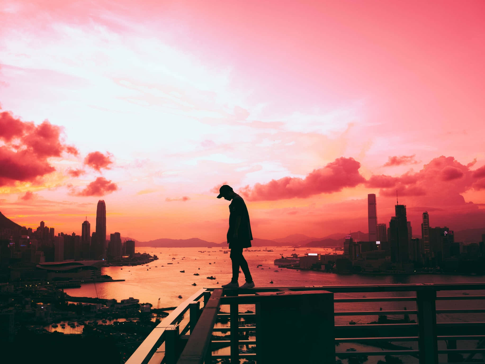 Pink Sky 桌布 壁紙 — 一個孤獨的身影站在天台欄杆上，俯瞰著沐浴在火熱粉紅和橙色夕陽色調中的城市景觀。