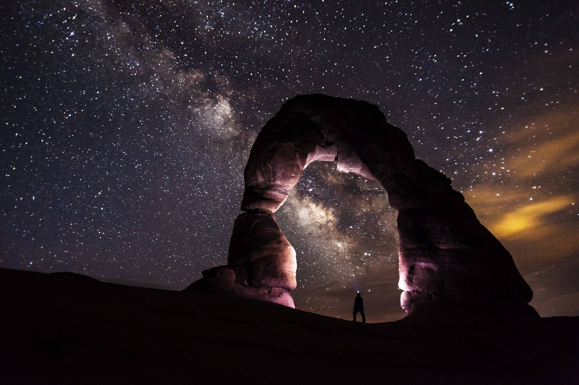 Pretty Galaxy 桌布 壁紙 — 一個孤獨的身影站在宏偉的石拱門下，拱門框住了銀河系發光的核心，背景是繁星點點的沙漠之夜。