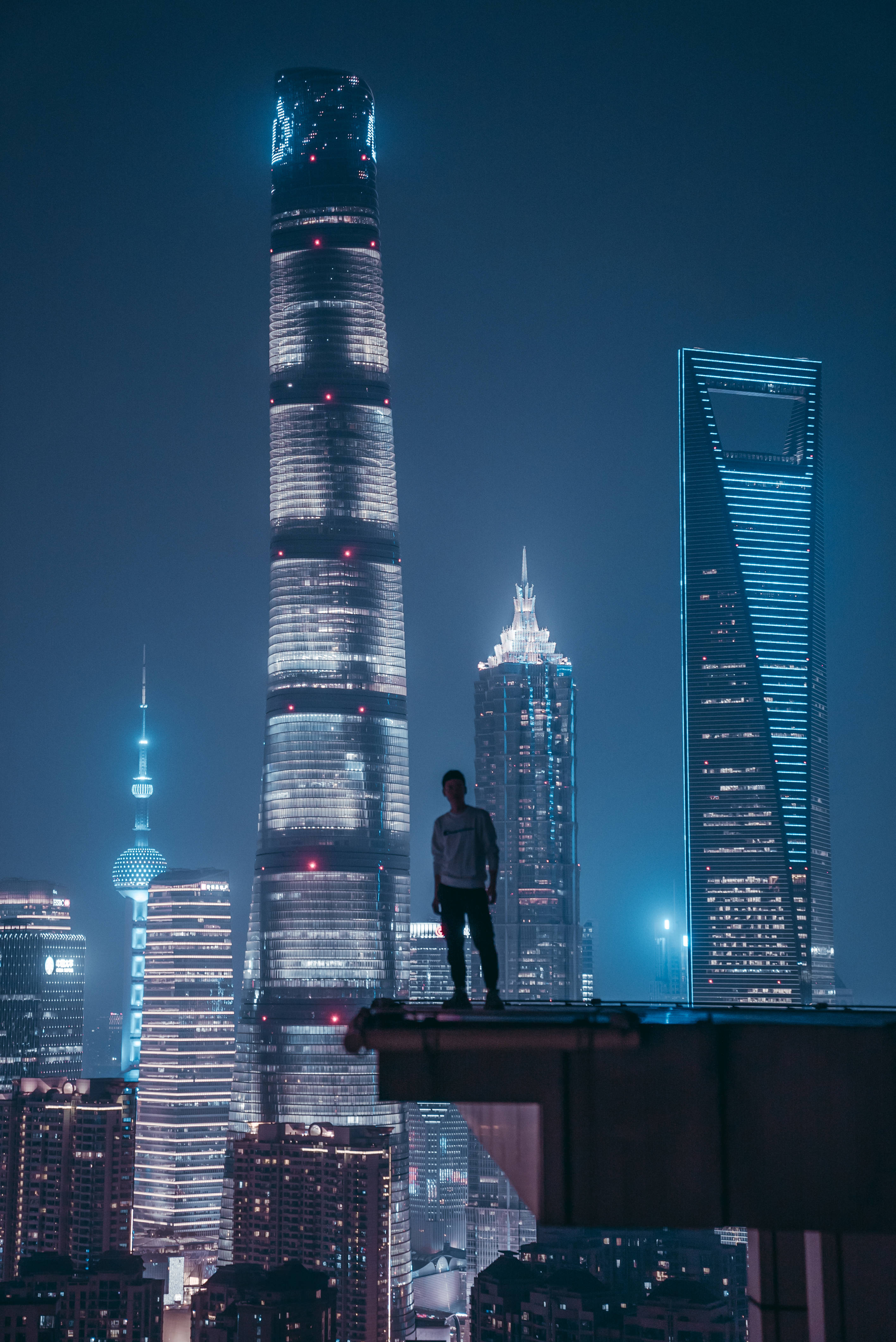Skyscraper 桌布 壁紙 — 一個孤獨的身影站在屋頂上，俯瞰上海燈火通明的摩天大樓，背景是深藍色的夜空。