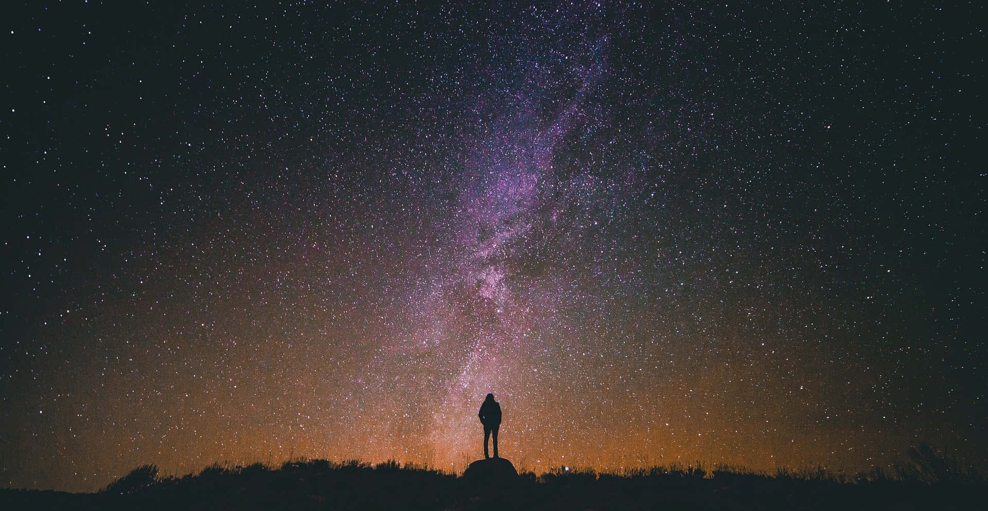 夜空 桌布 壁紙 — 一個孤獨的身影站在山頂上，仰望充滿銀河系的壯麗紫色和橙色星空。